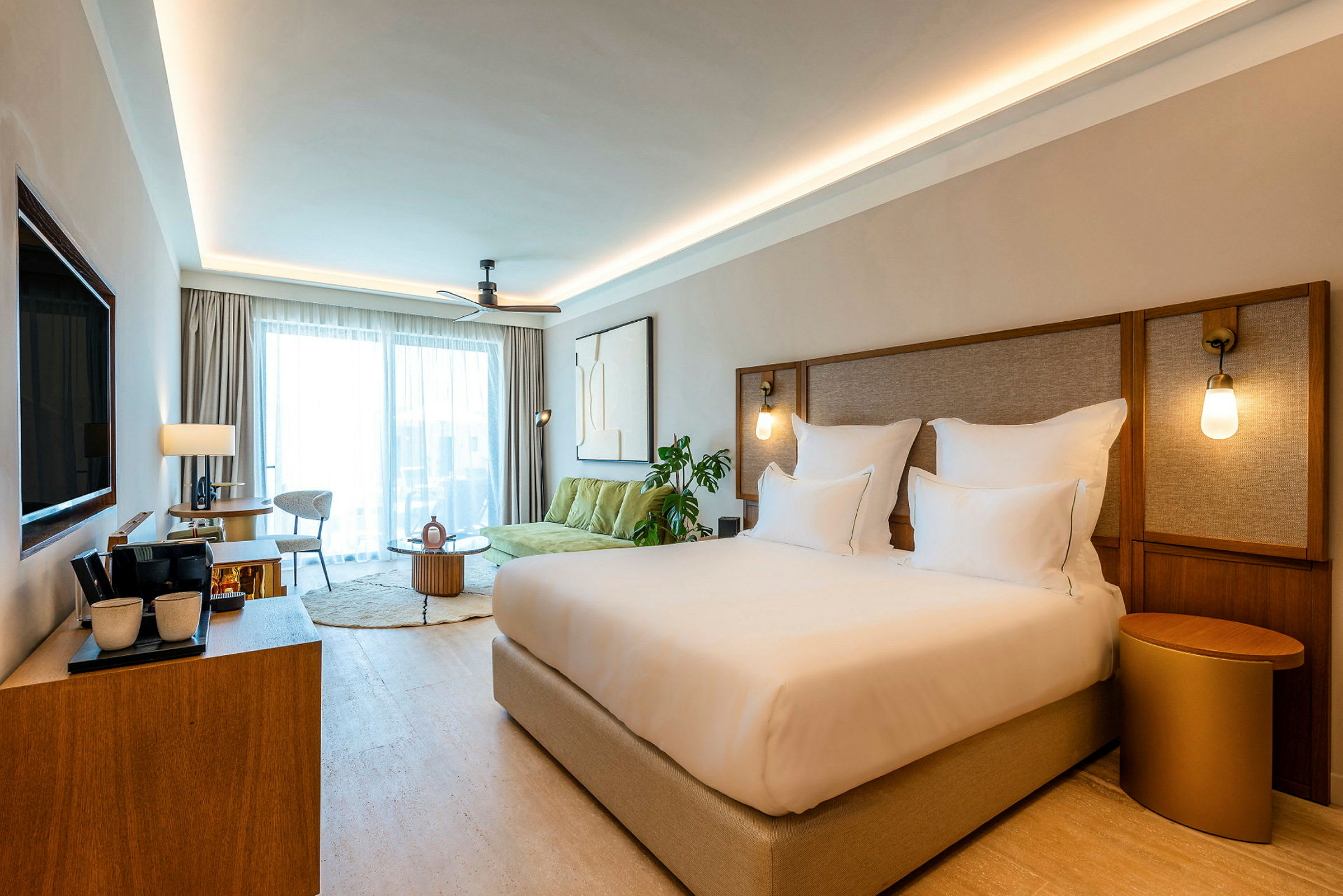 Et elegant hotellrom med en stor seng, sittegruppe og store vinduer med gardiner. Vegglamper gir varm belysning. (AI-generert tekst)