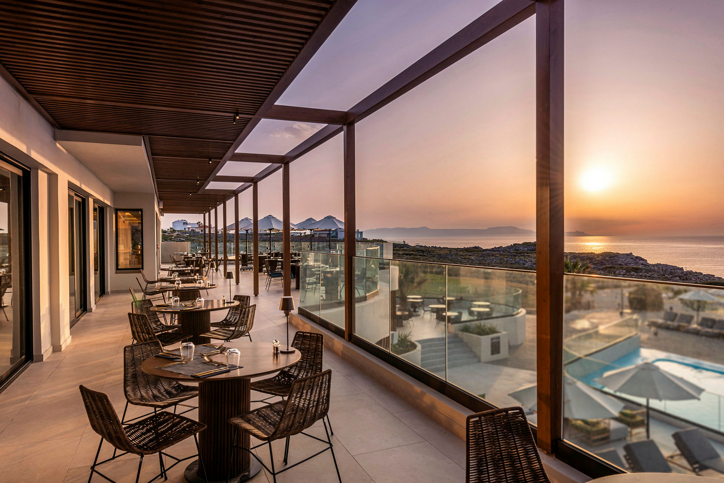 En terrasse med bord og stoler, omgitt av glassrekkverk, med utsikt over havet og solnedgangen. (AI-generert tekst)