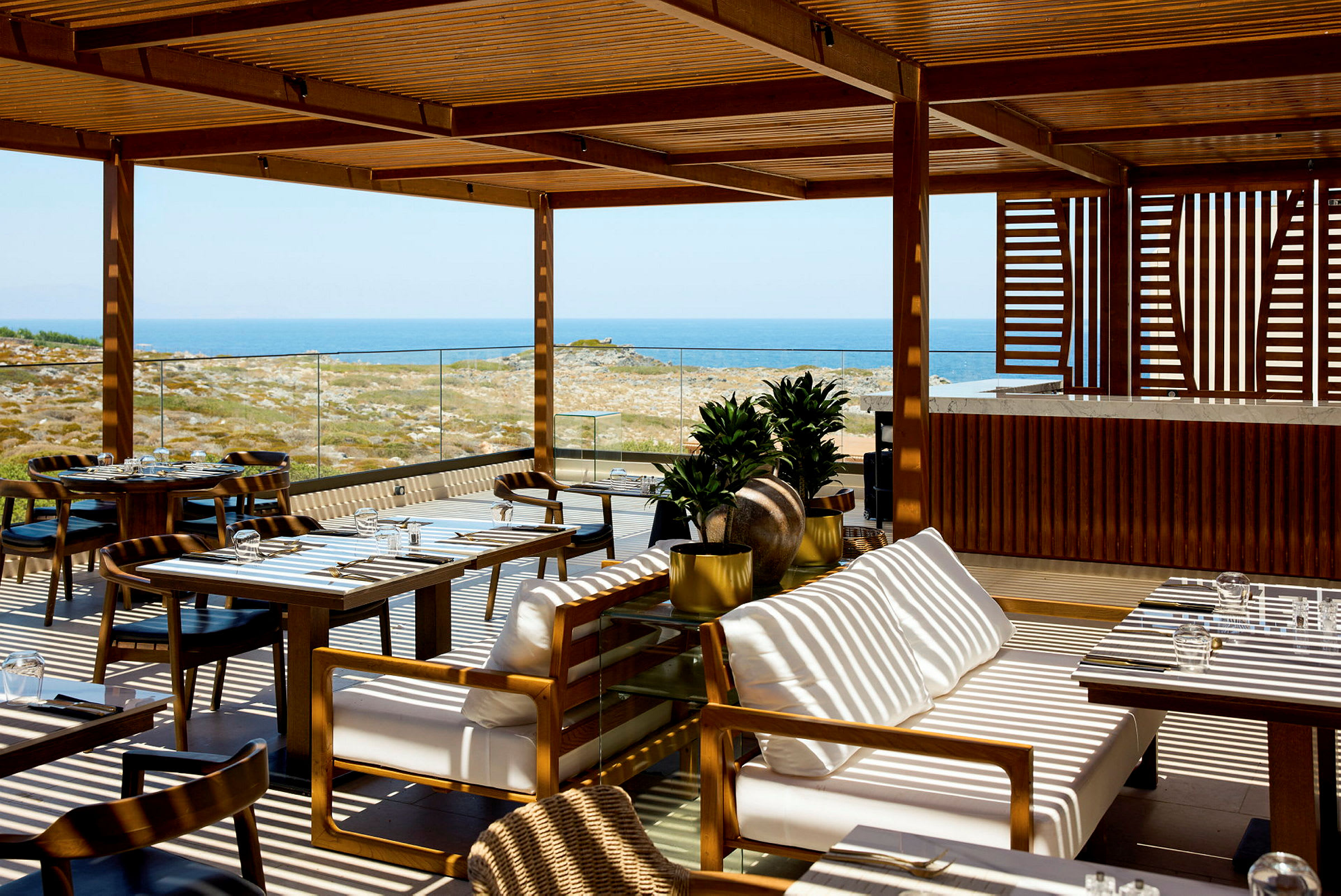 En utendørsrestaurant med trepergola, bord og stoler samt havutsikt i bakgrunnen. (AI-generert tekst)