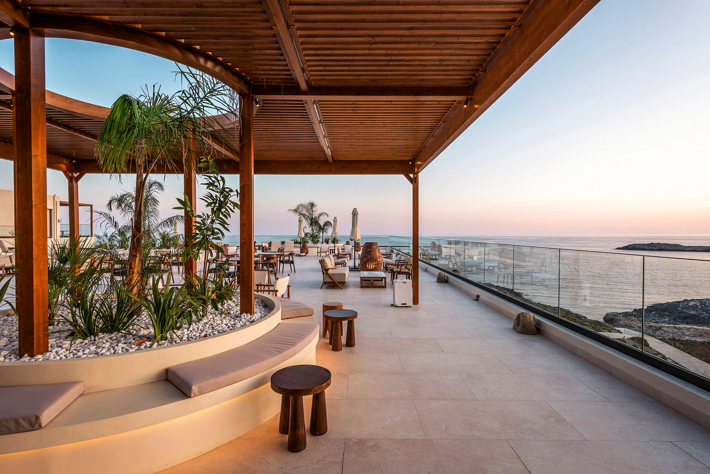 Utendørsområde med sitteplasser under en trepergola, omgitt av planter og med utsikt over havet. (AI-generert tekst)