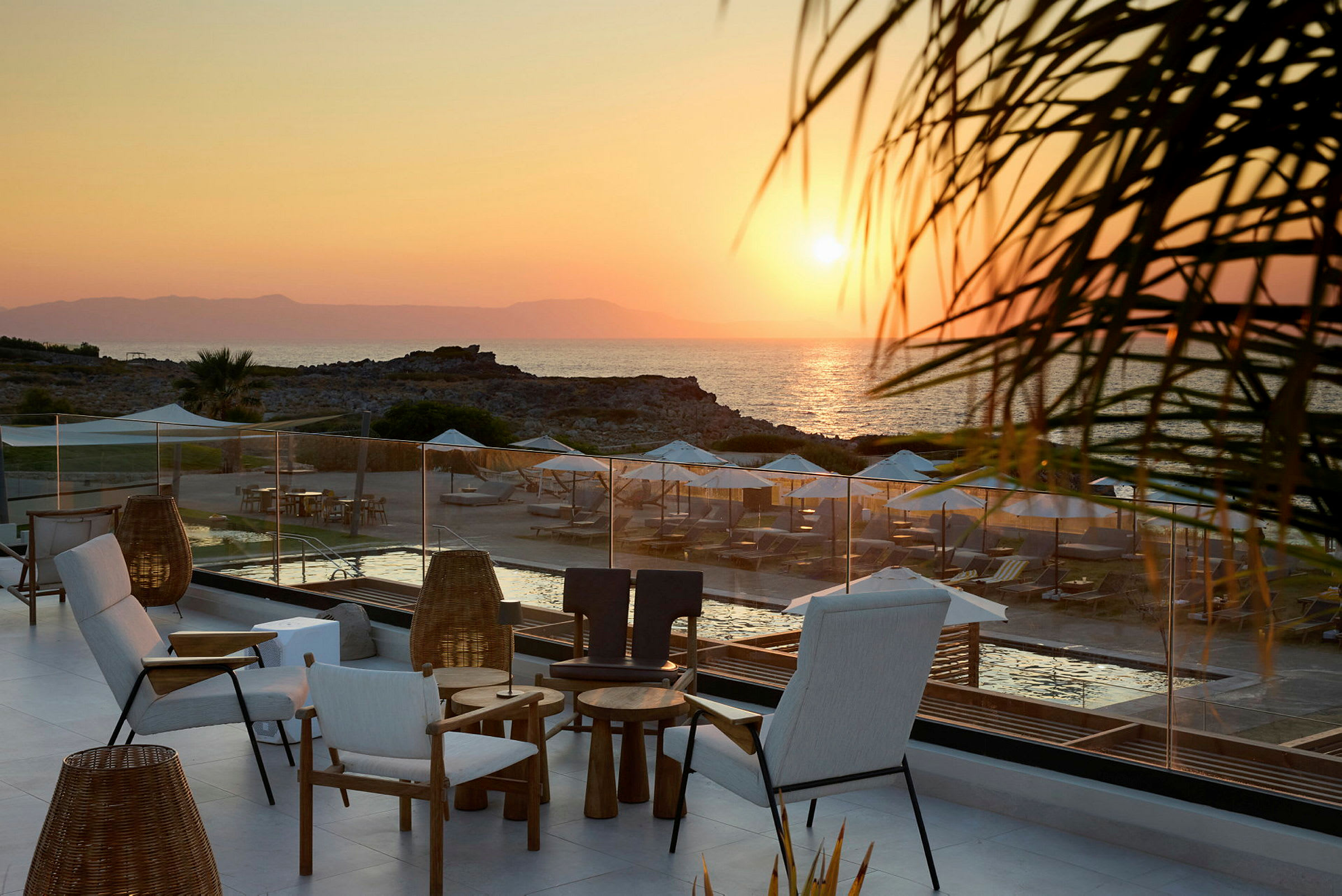 Terrasse med sitteplasser i forgrunnen, med utsikt over en strand med solsenger og parasoller ved solnedgang. (AI-generert tekst)