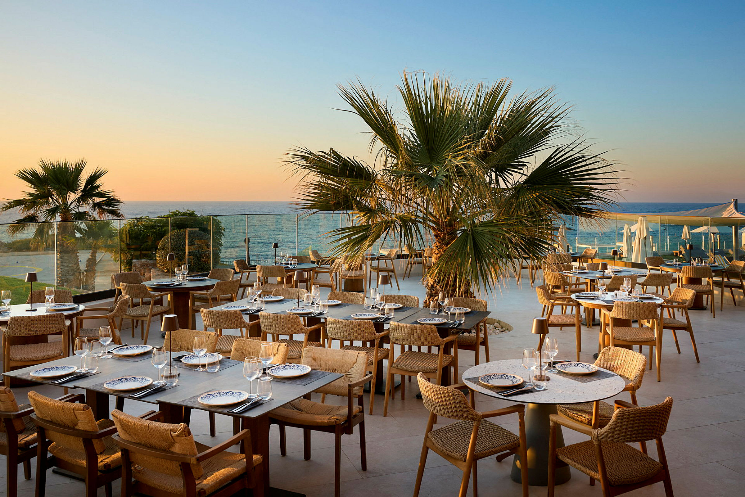 En utendørs restaurant med bord og stoler, omgitt av palmer og med utsikt over havet i solnedgangen. (AI-generert tekst)