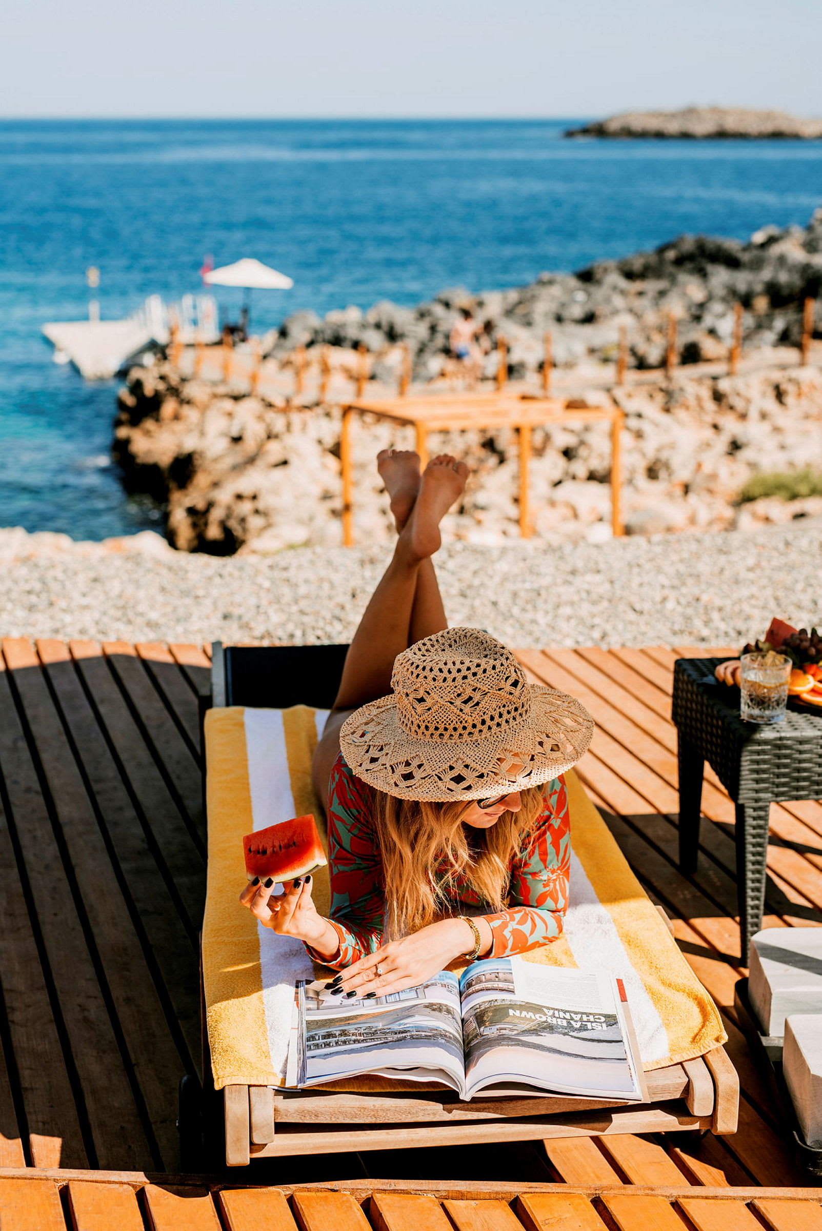 En person ligger på en solseng på en solfylt dag med havet og klipper i bakgrunnen. Personen holder et stykke vannmelon og leser en bok. (AI-generert tekst)