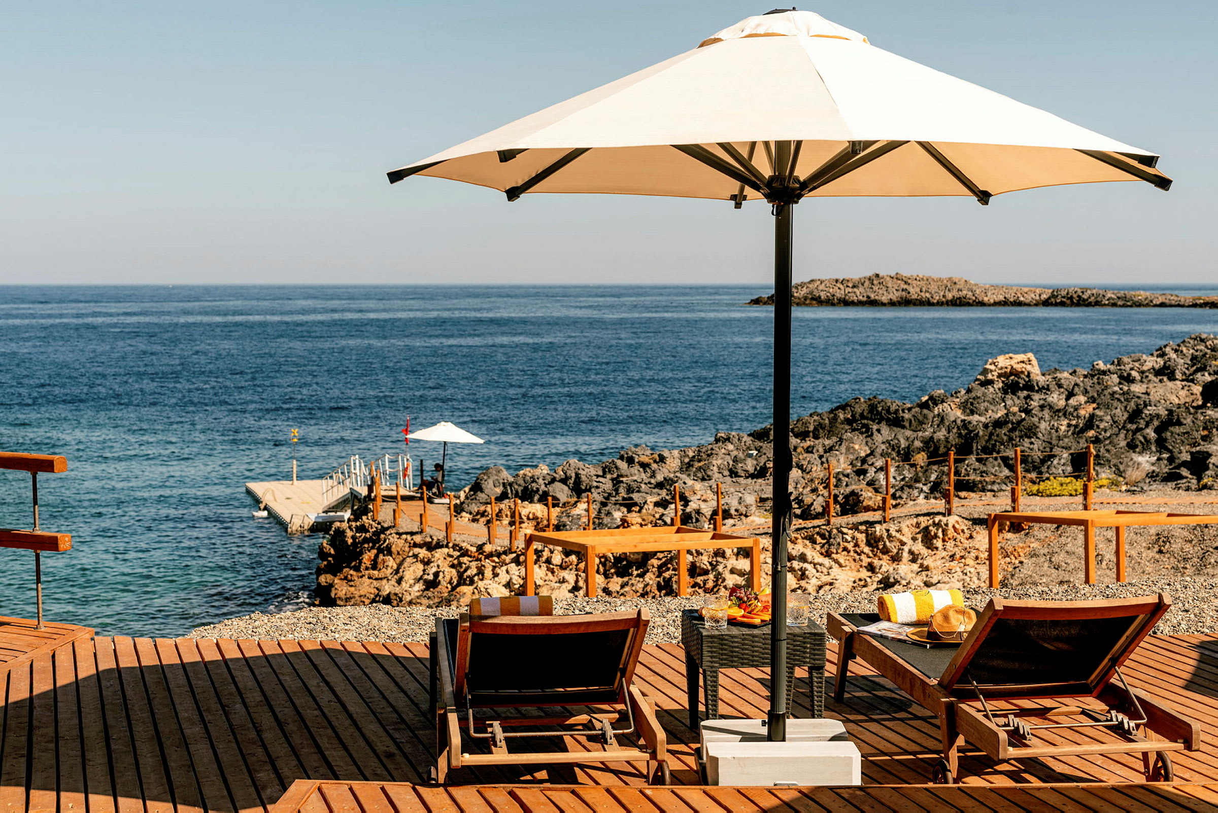 Et loungeområde nær havet med to solsenger, en parasoll og et bord med drikker og frukt. I bakgrunnen er det klipper og en liten brygge. (AI-generert tekst)