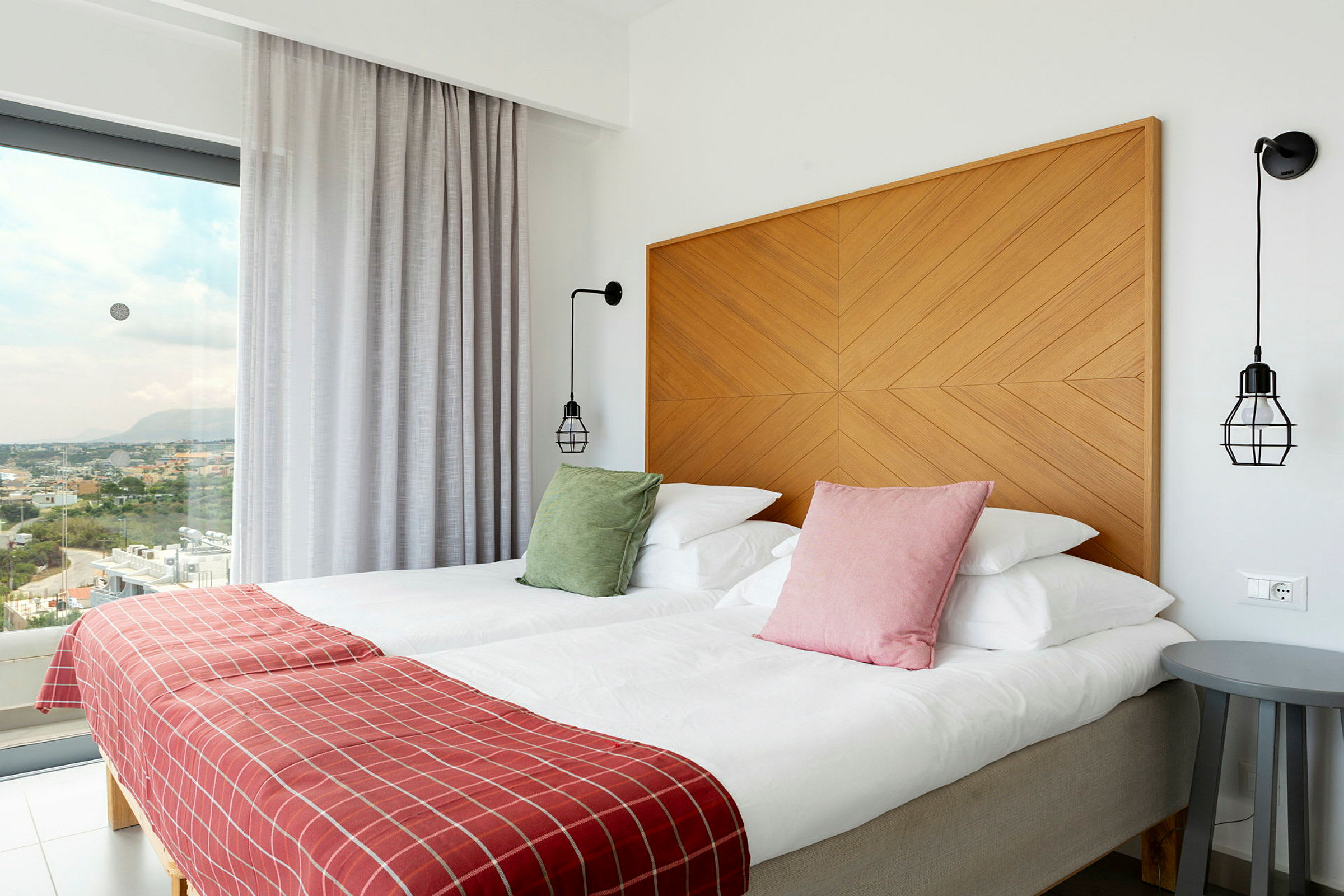 Hotellrom med to senger, fargerike puter, rødt sengeteppe og moderne innredning, med utsikt gjennom store vinduer. (AI-generert tekst)