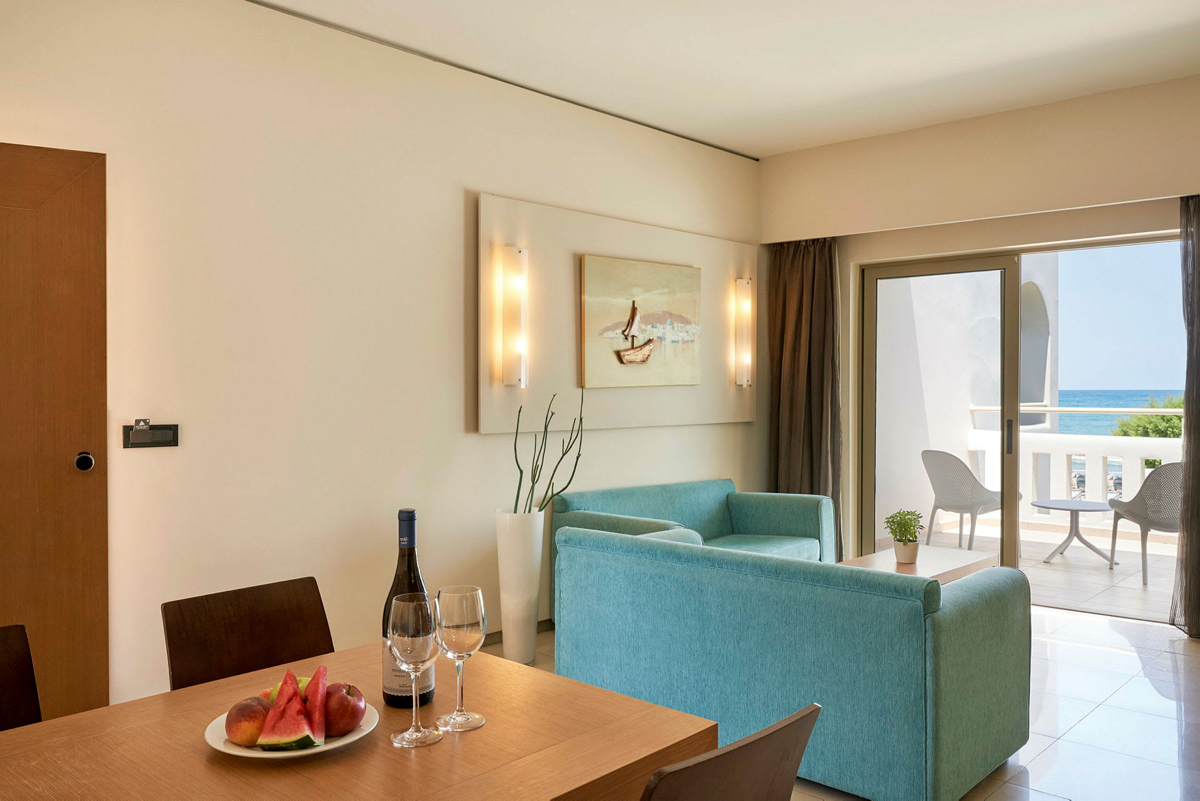Et hotellrom med spiseplass og sittegruppe, samt en balkong med havutsikt. (AI-generert tekst)
