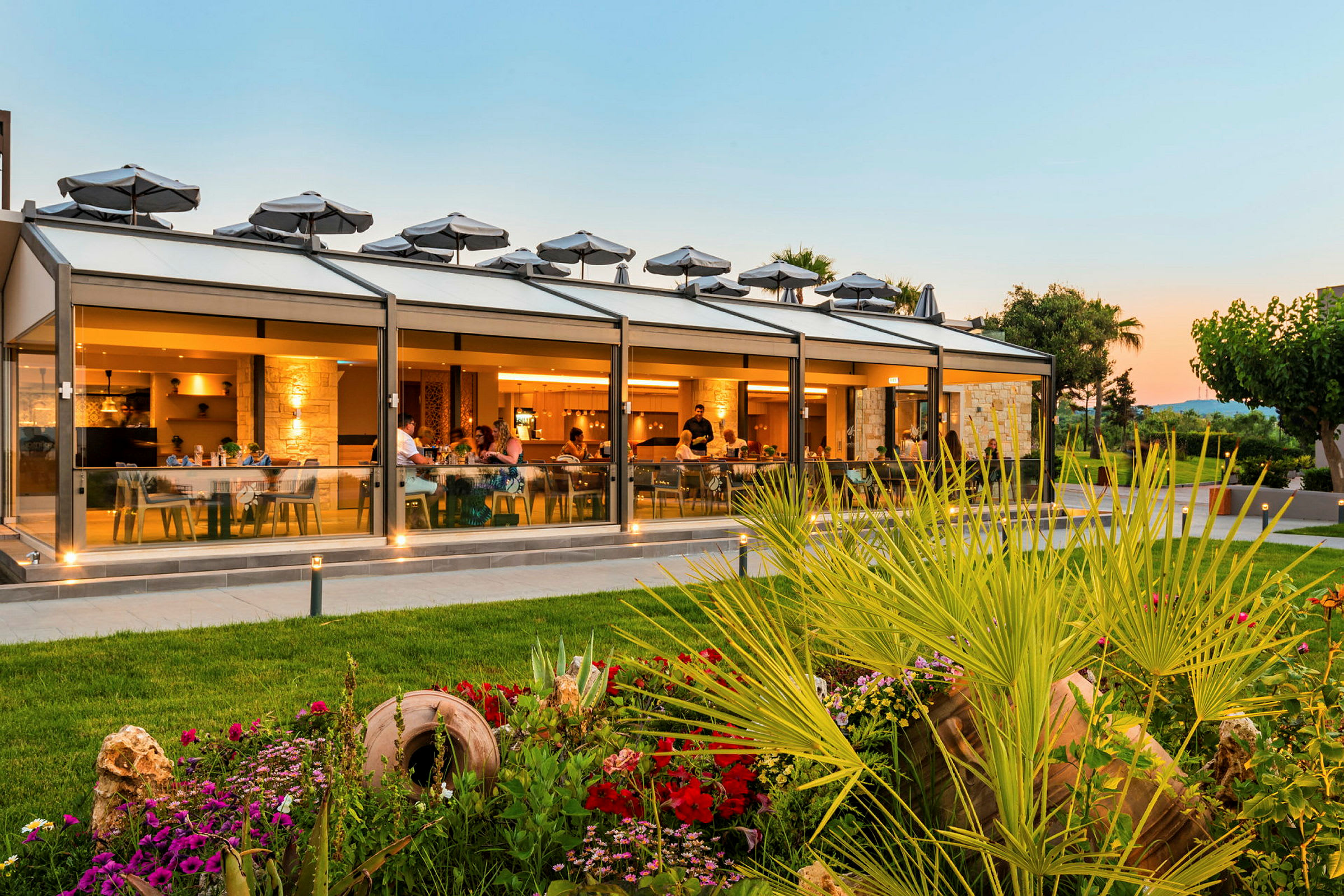 En utendørs restaurant med glassvegger, omgitt av en hage med planter og blomster. (AI-generert tekst)