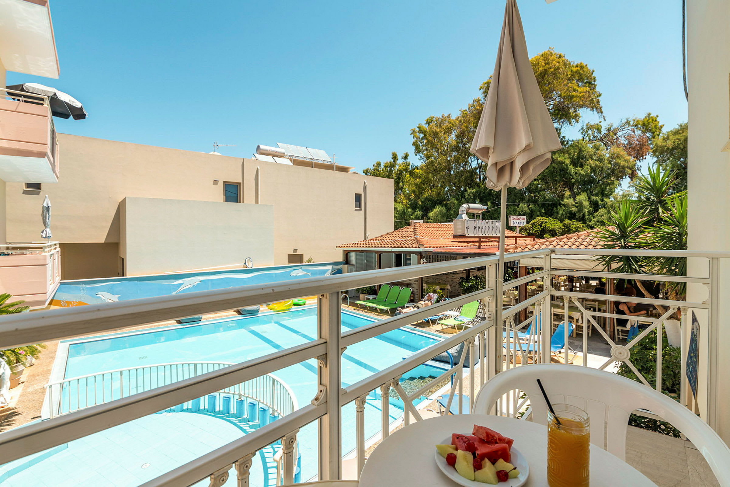 Utsikt fra en balkong mot et basseng og hotellområde med solsenger, parasoller og grøntområder. På balkongen står et bord med en tallerken melon og et glass juice. (AI-generert tekst)