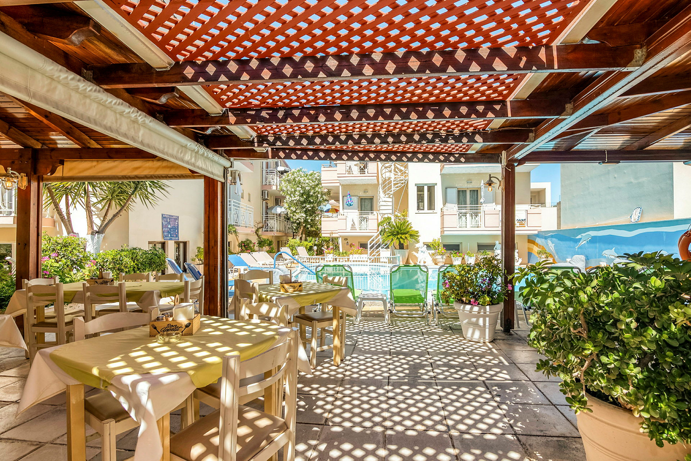 Bilde av et utendørs spiseområde med bord og stoler under en trepergola, med utsikt mot et basseng og bygninger i bakgrunnen. (AI-generert tekst)