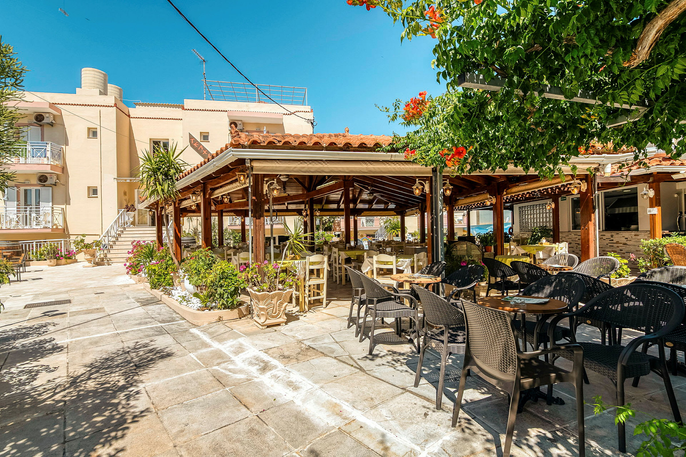 En utendørsrestaurant med bord og stoler under en pergola, omgitt av grønne planter. (AI-generert tekst)