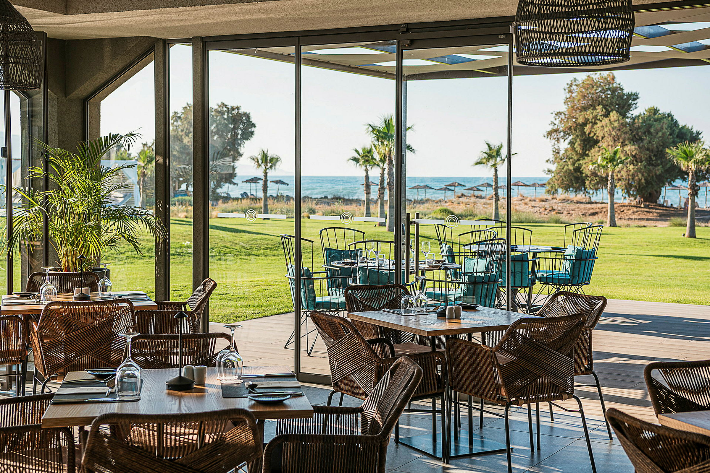 En restaurant med glassvegger og utsikt over en grønn plen, palmer og havet. (AI-generert tekst)