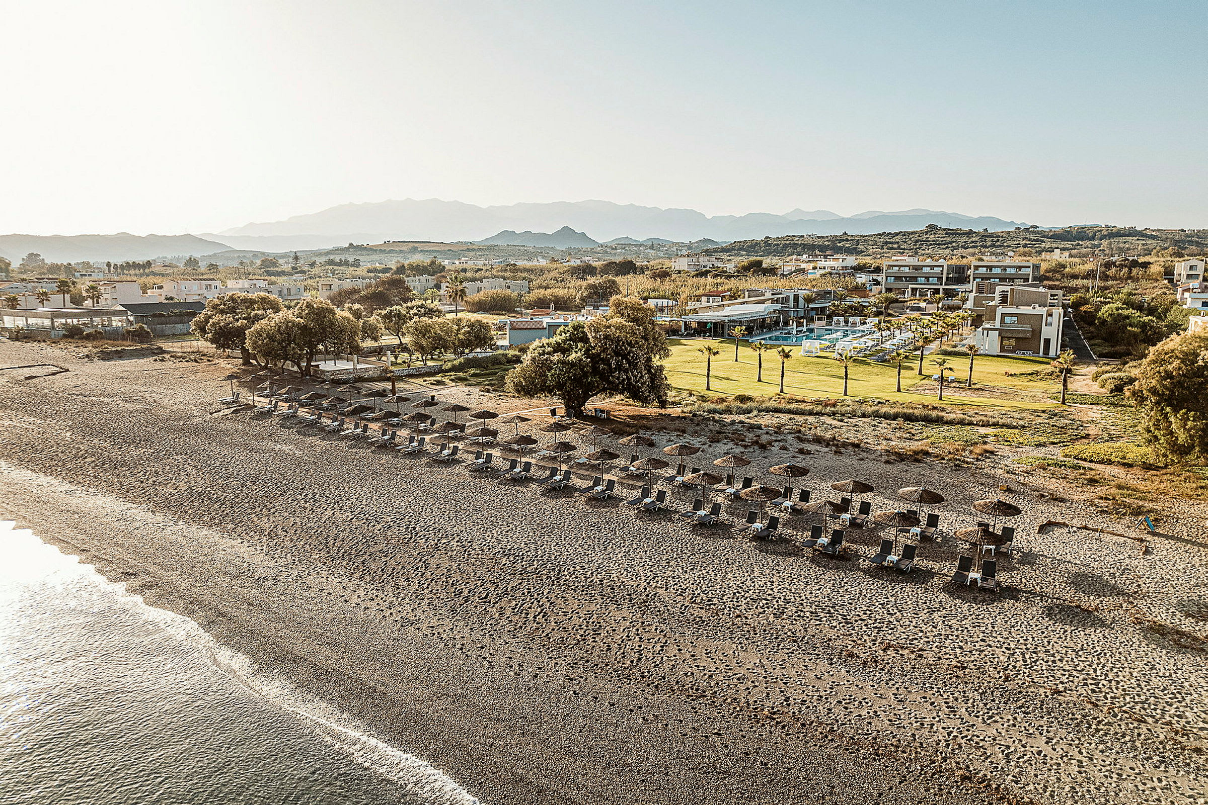 En strand med solsenger og parasoller, omgitt av grønne områder og hotellbygninger i bakgrunnen. (AI-generert tekst)