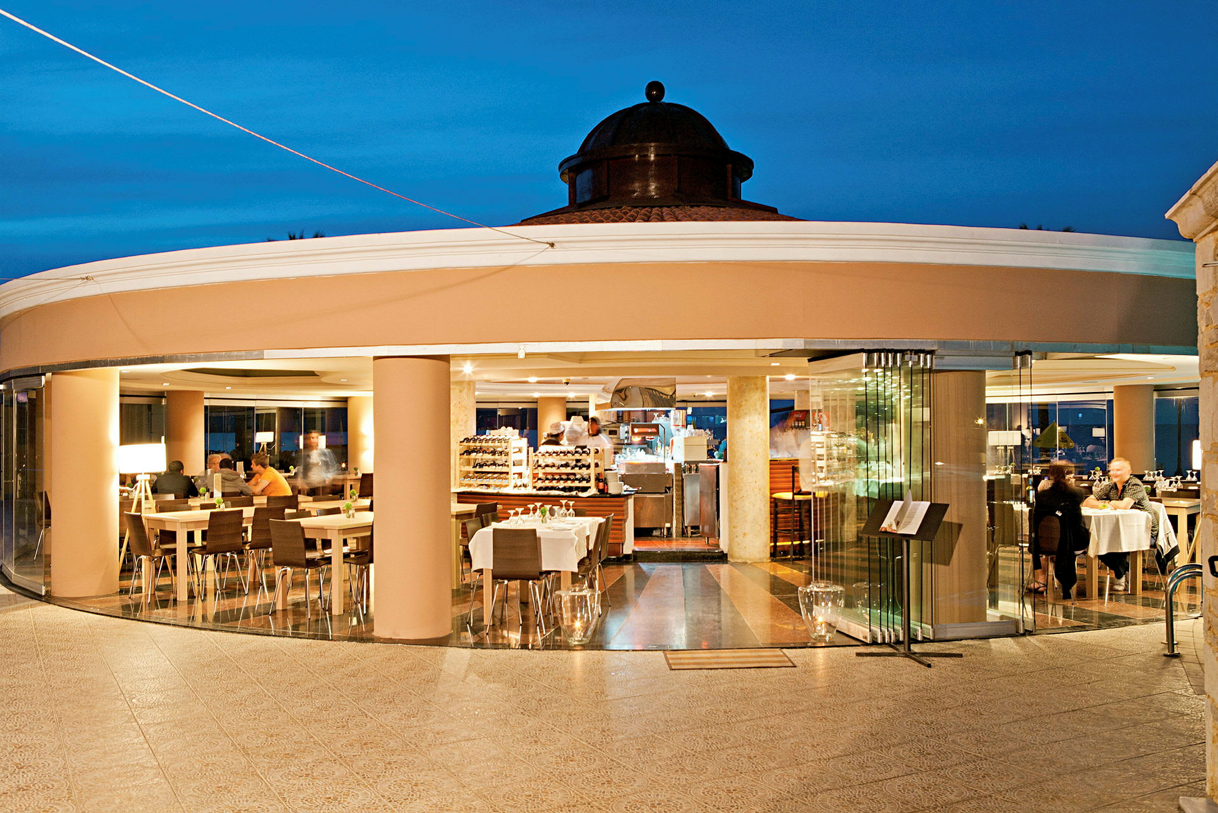 En restaurant med glassvegger og bord plassert innendørs, sett fra en utendørsterrasse. (AI-generert tekst)