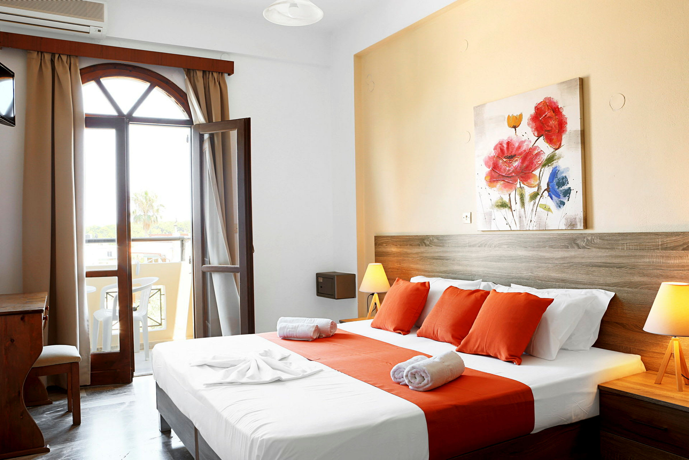 Et hotellrom med en seng kledd i hvite og oransje laken, et tre-skrivebord og en åpning mot en balkong. (AI-generert tekst)