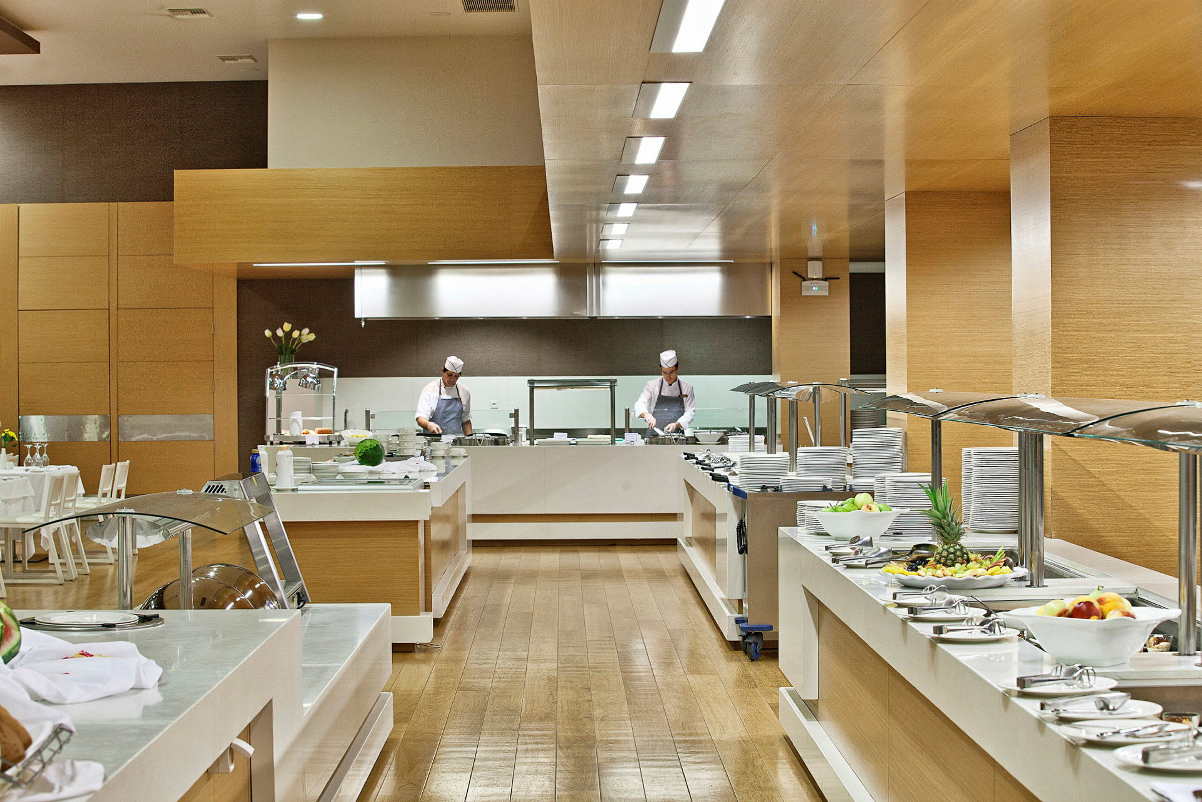 Et bufféområde i en moderne restaurant med tallerkener, bestikk og mat oppstilt, samt personale som forbereder mat. (AI-generert tekst)