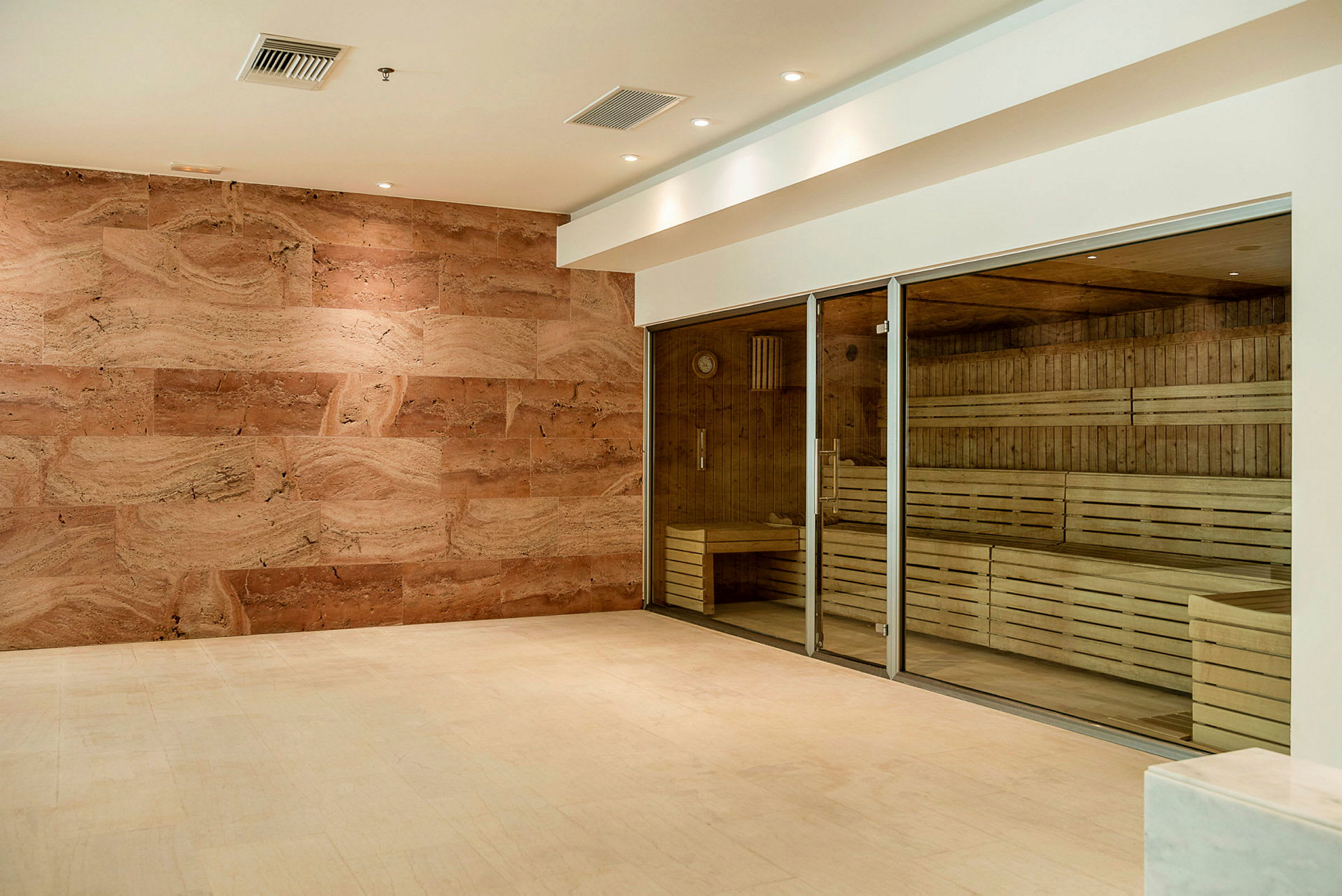 En badstue med glassdører og trebenker er synlig, omgitt av et rom med sandfargede vegger og gulv. (AI-generert tekst)