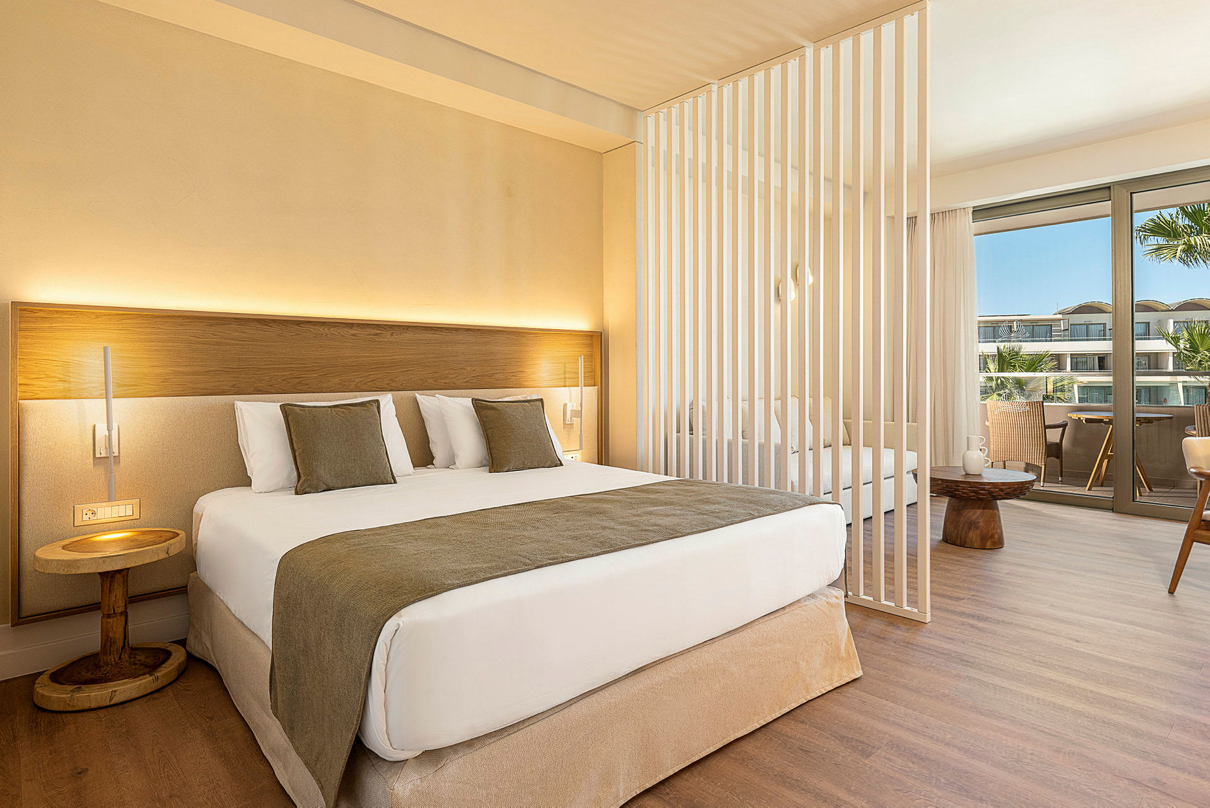 Et moderne hotellrom med en dobbeltseng, dekorative puter og et lite nattbord. Rommet har en skillevegg og en sittegruppe med utsikt over en balkong. (AI-generert tekst)