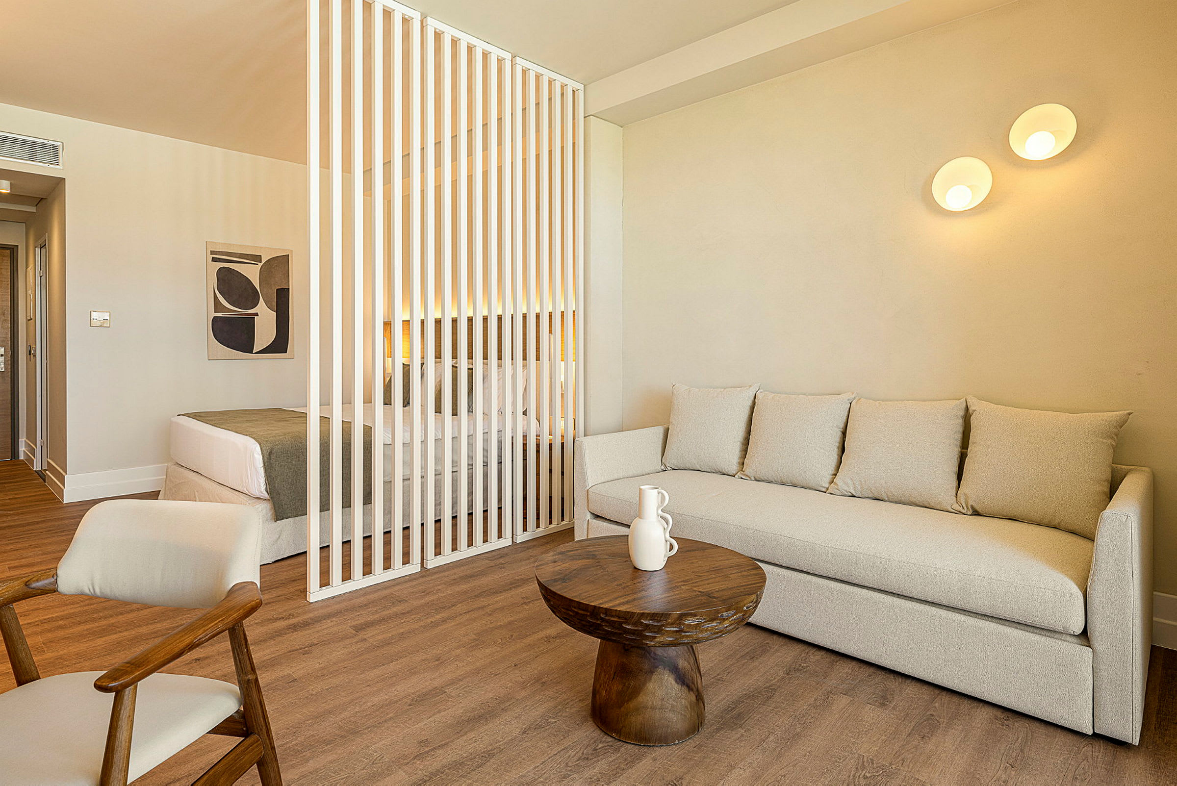 Et rom med en sittegruppe bestående av en beige sofa og et lite rundt trebord foran. I bakgrunnen er det en dobbeltseng adskilt med hvite vertikale ribber. (AI-generert tekst)