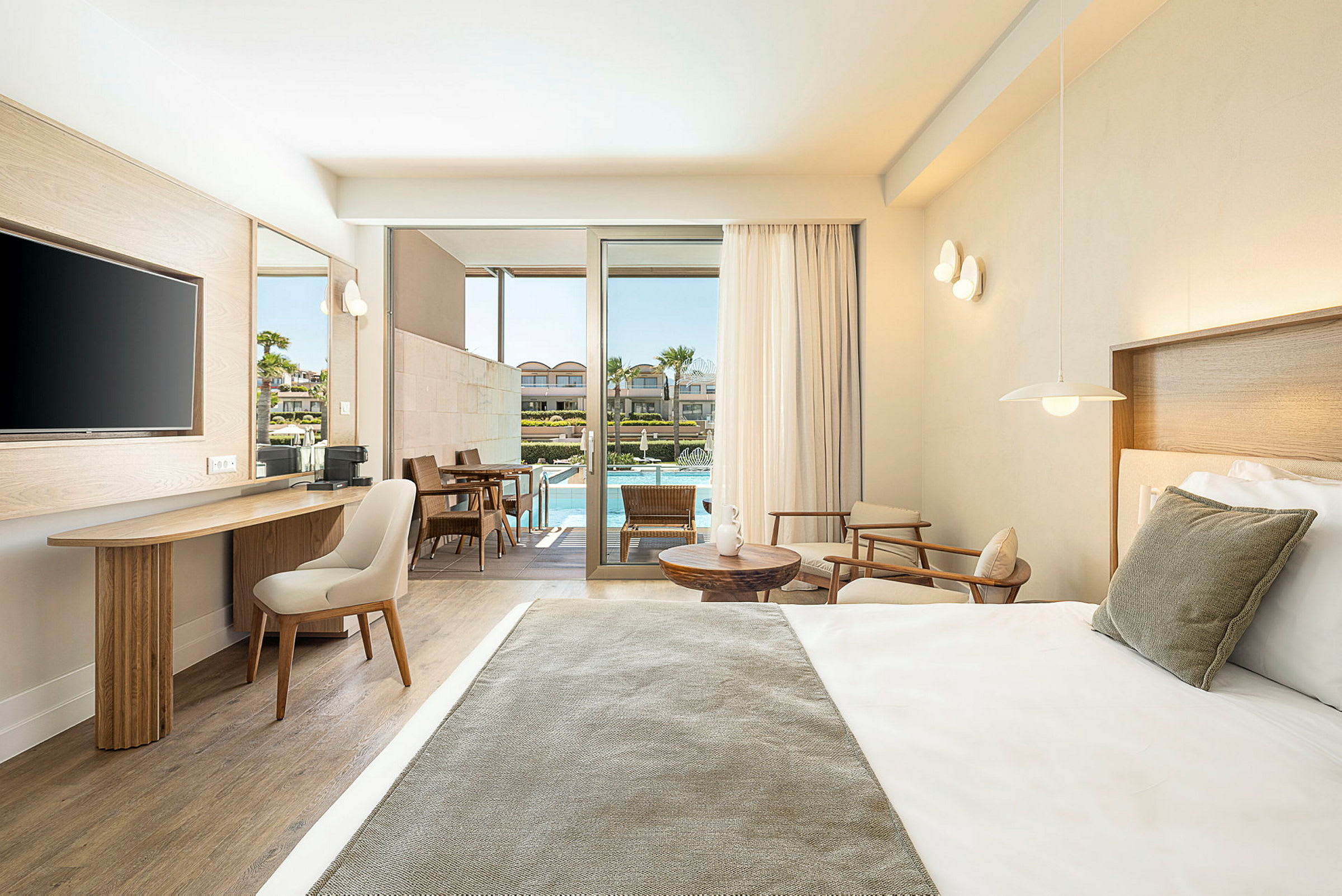 Et moderne hotellrom med en oppredd seng, sittegruppe og skrivebord. Rommet har store glassdører ut til en terrasse med bord, stoler og tilgang til et svømmebasseng. (AI-generert tekst)
