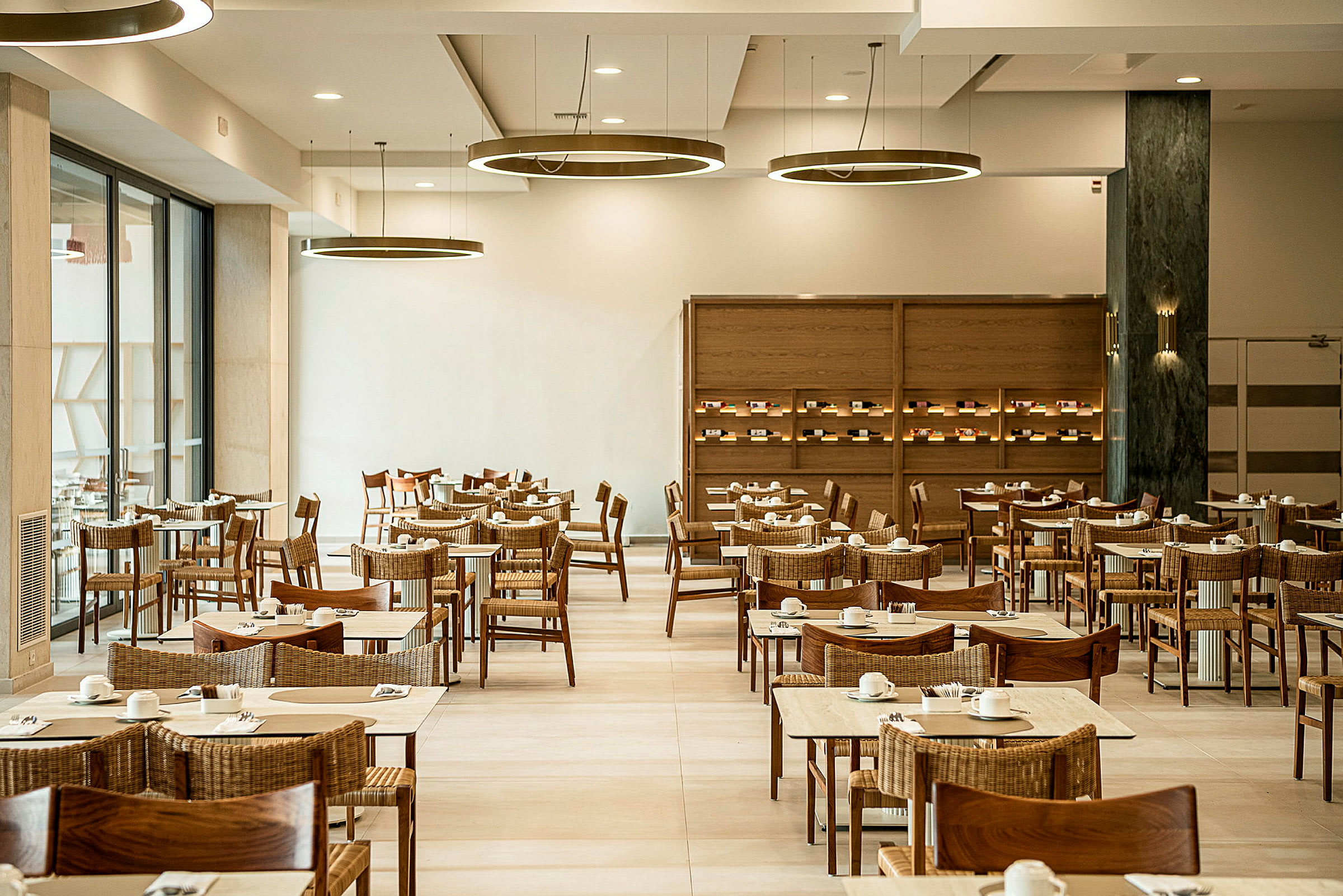 Et spiseområde med flere trebord og stoler, dekket med kopper og servietter. Rommet har moderne belysning og naturlig lysinnslipp fra store vinduer. (AI-generert tekst)