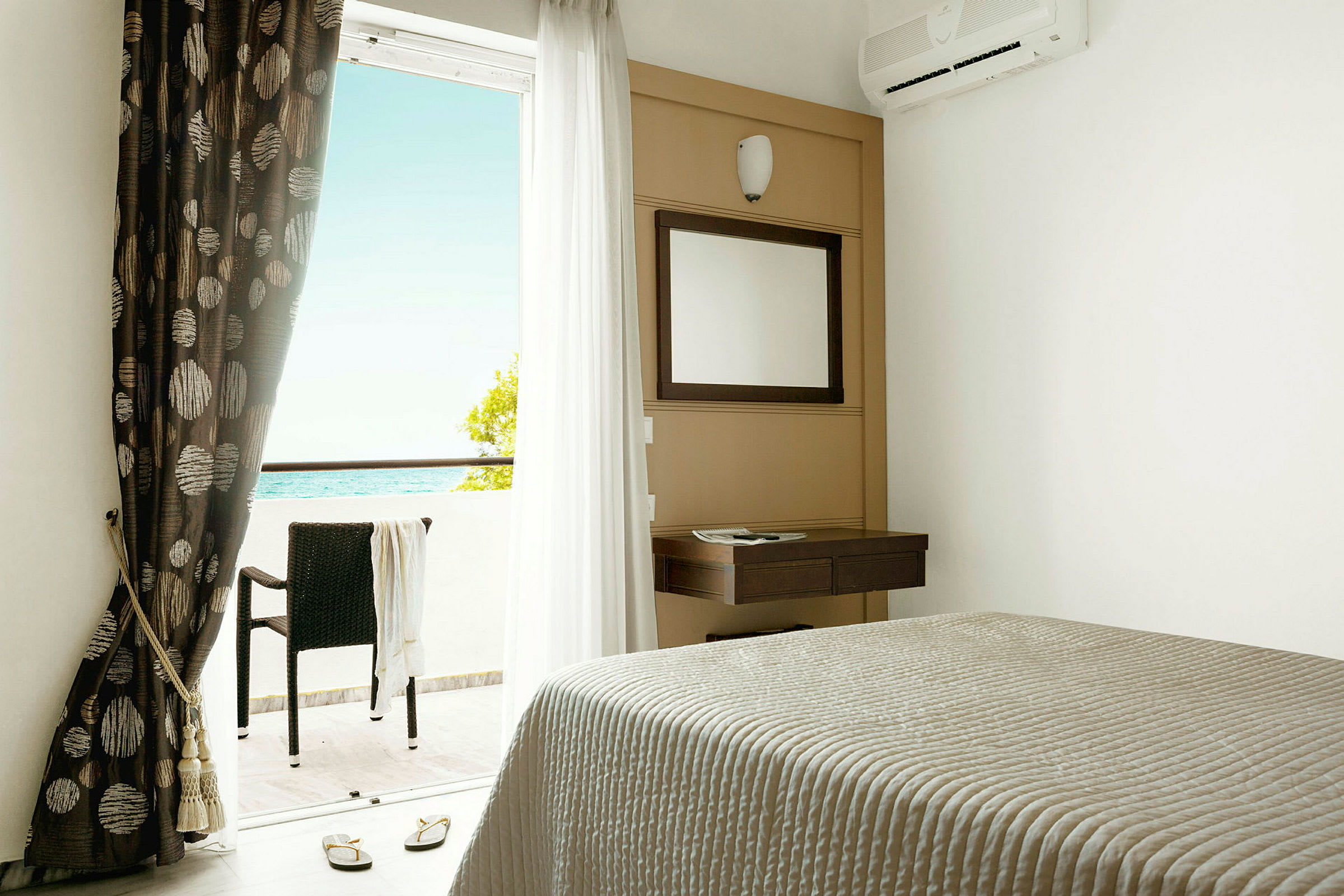 Hotellrom med seng, speil, gardiner og balkong med utsikt mot havet. (AI-generert tekst)