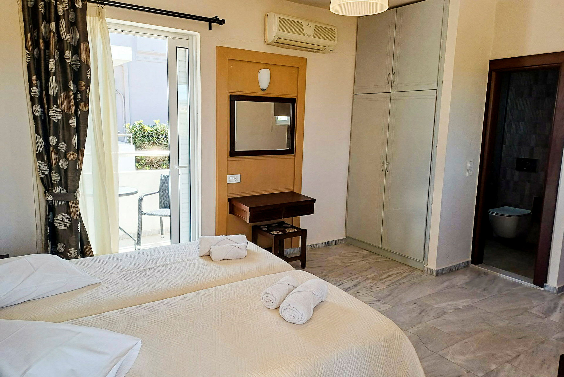 Hotellrom med to enkeltsenger, et skrivebord og utsikt mot en balkong med sitteplass. (AI-generert tekst)