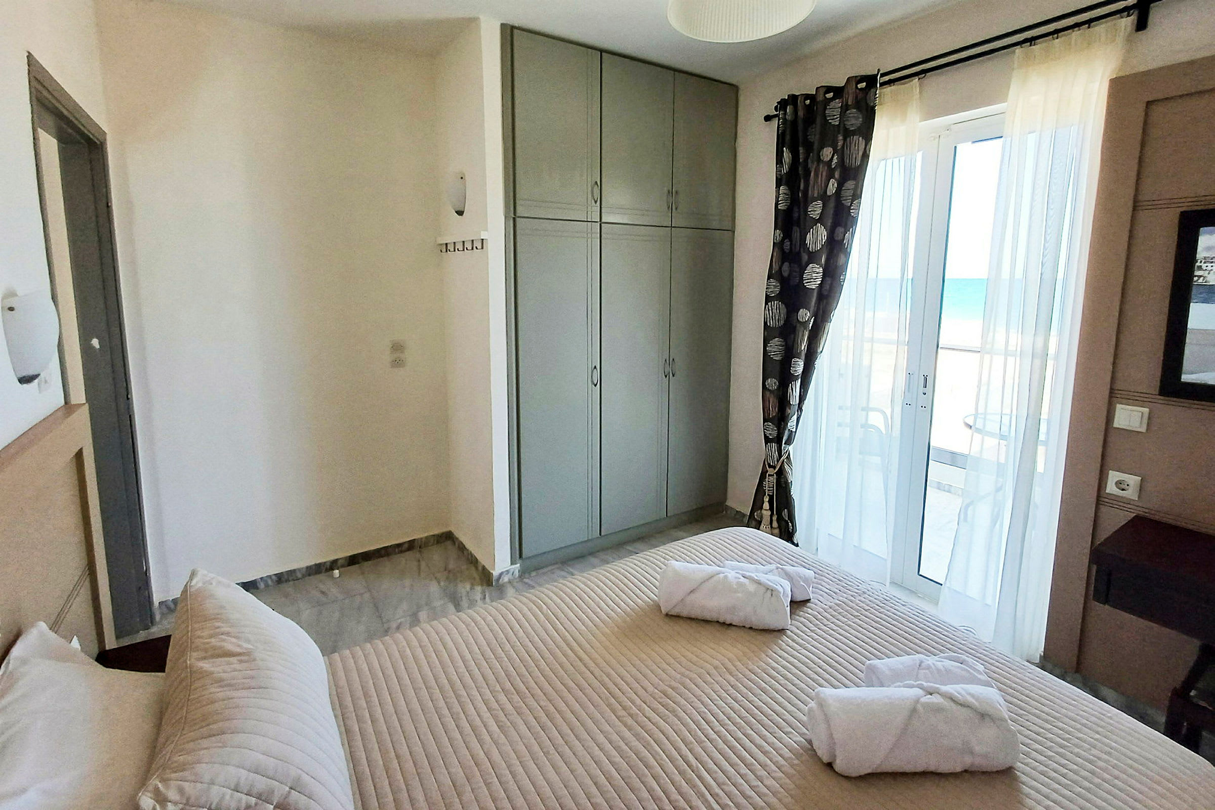 Et hotellrom med en seng, grå garderobe og en balkongdør med havutsikt. (AI-generert tekst)