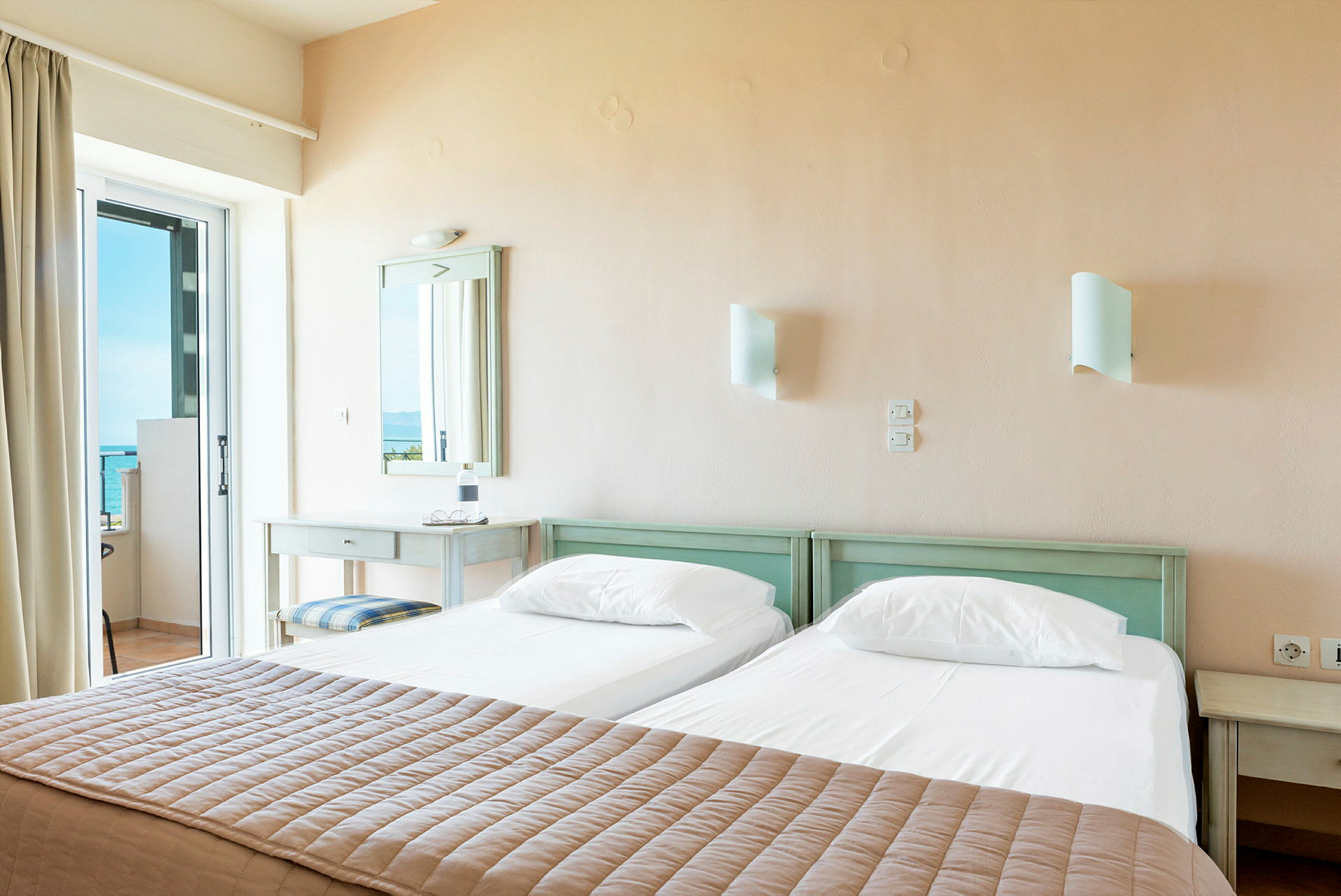 Et hotellrom med to enkeltsenger og utsikt over havet gjennom en glassdør. (AI-generert tekst)