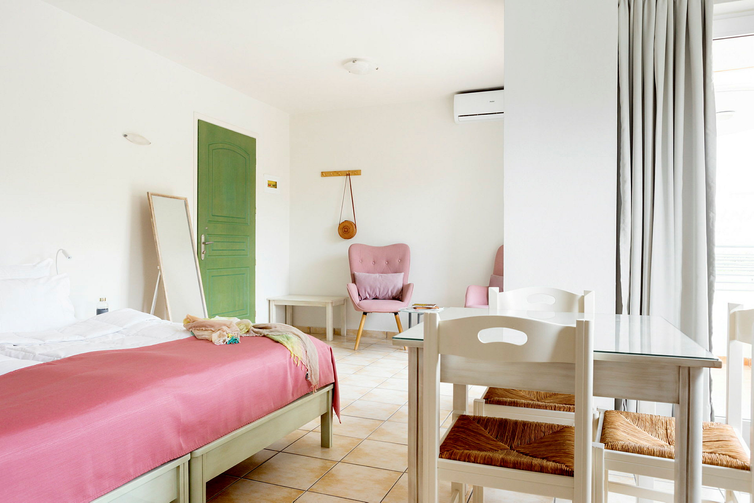 Et lyst og romslig hotellrom med en grønn dør, en seng med rosa overkast, og et bord med stoler. (AI-generert tekst)