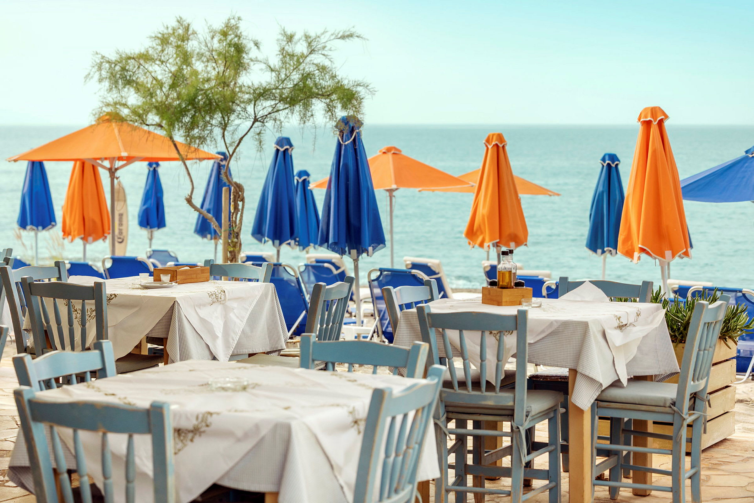 Et bord og stoler står på en terrasse med utsikt over havet. I bakgrunnen kan man se parasoller i oransje og blått på stranden. (AI-generert tekst)