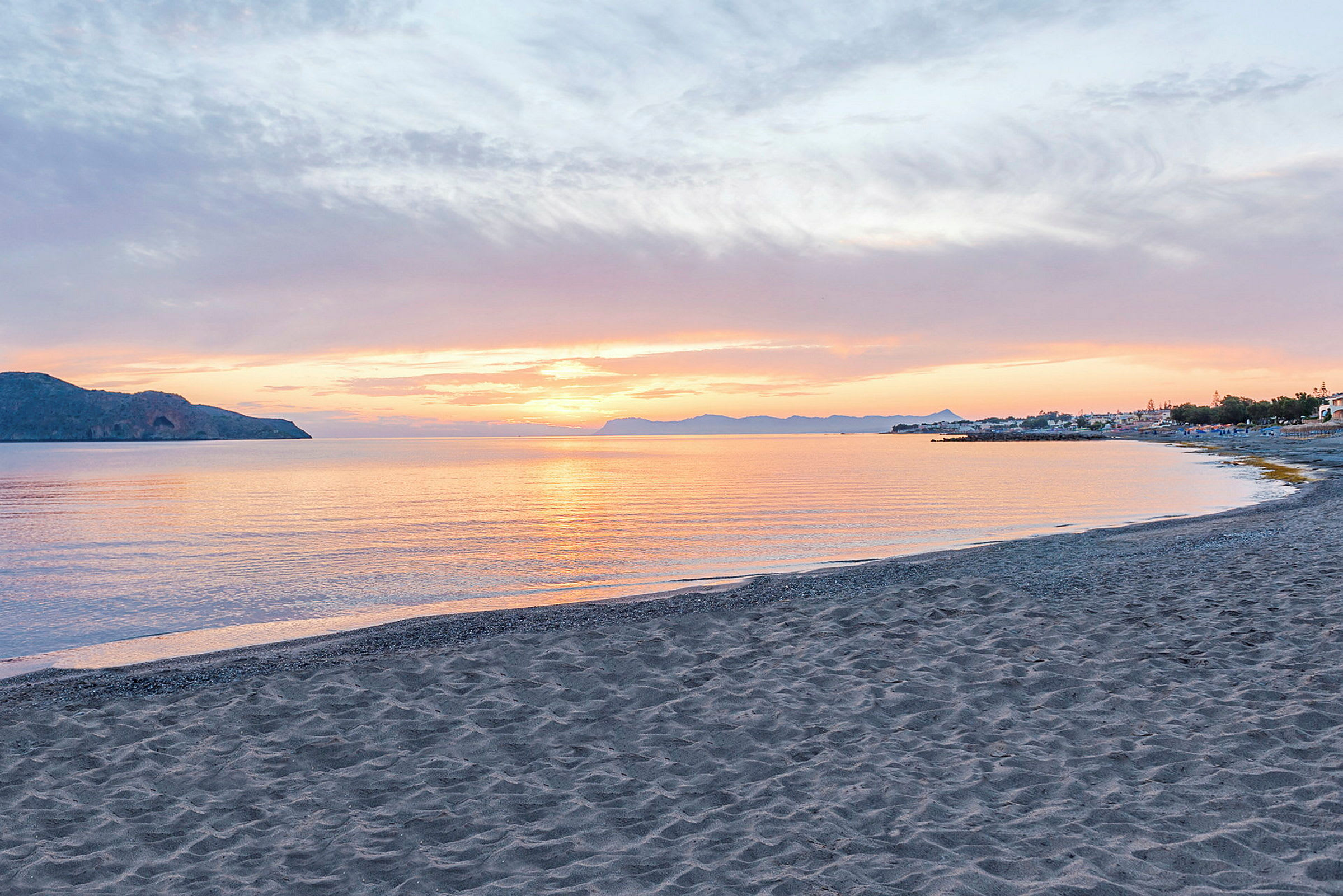 Solnedgang sett fra en strand med sjøen i forgrunnen og fjell og bygninger i bakgrunnen. (AI-generert tekst)