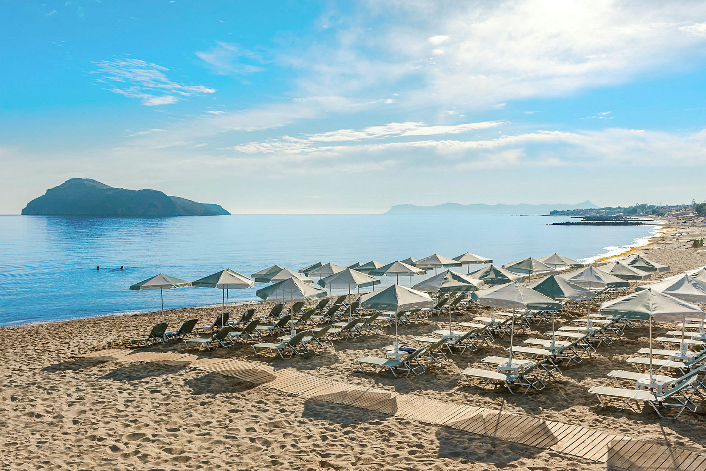 Utsikt over en strand med solsenger og parasoller, omgitt av hav og en fjern øy. (AI-generert tekst)