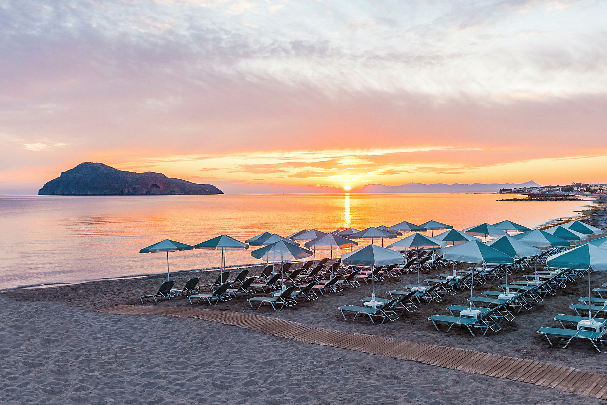 Solnedgang på en sandstrand med parasoller og solsenger, i bakgrunnen en øy og rolig hav. (AI-generert tekst)