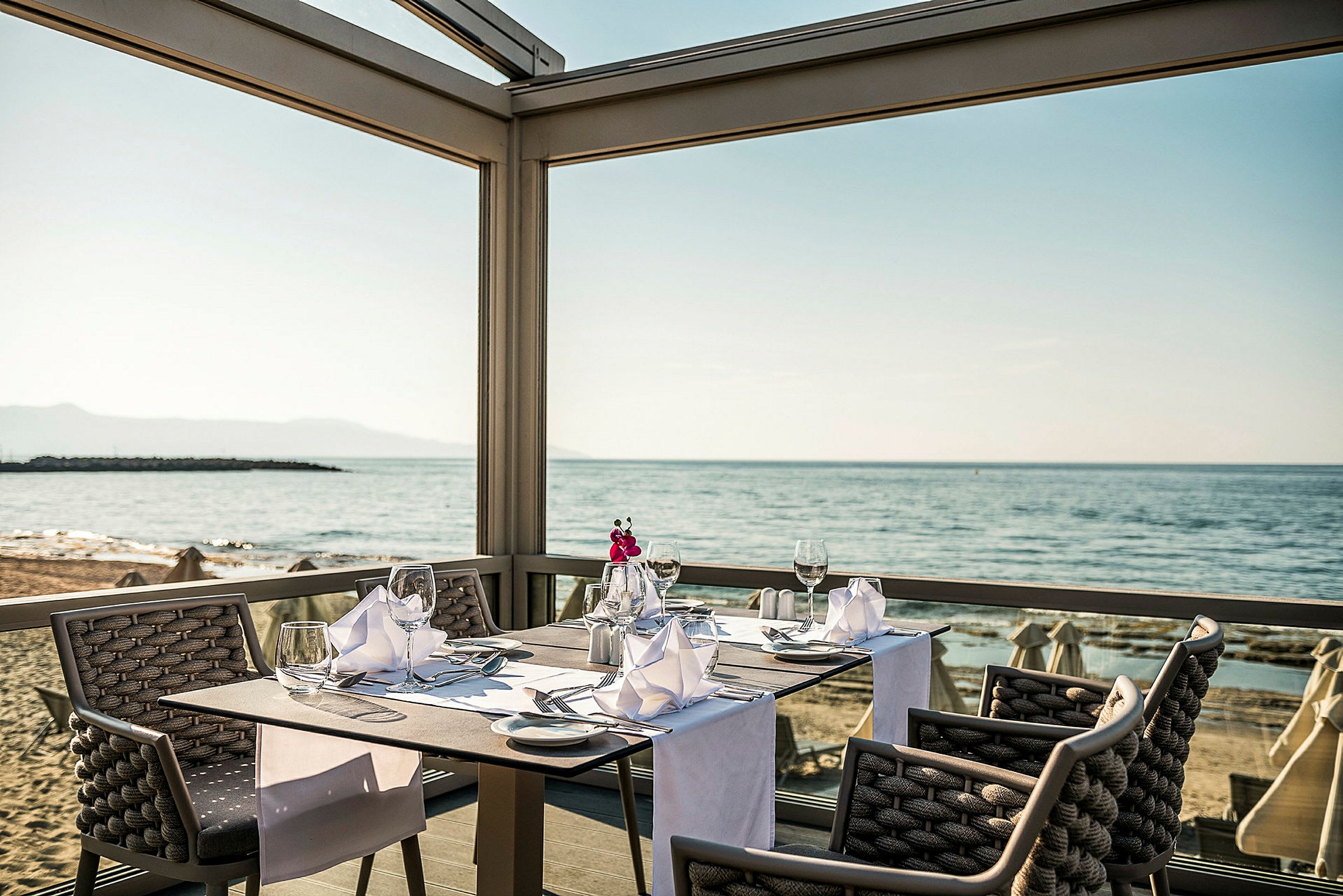 En restaurantplass ved sjøen med dekket bord og utsikt over stranden. (AI-generert tekst)