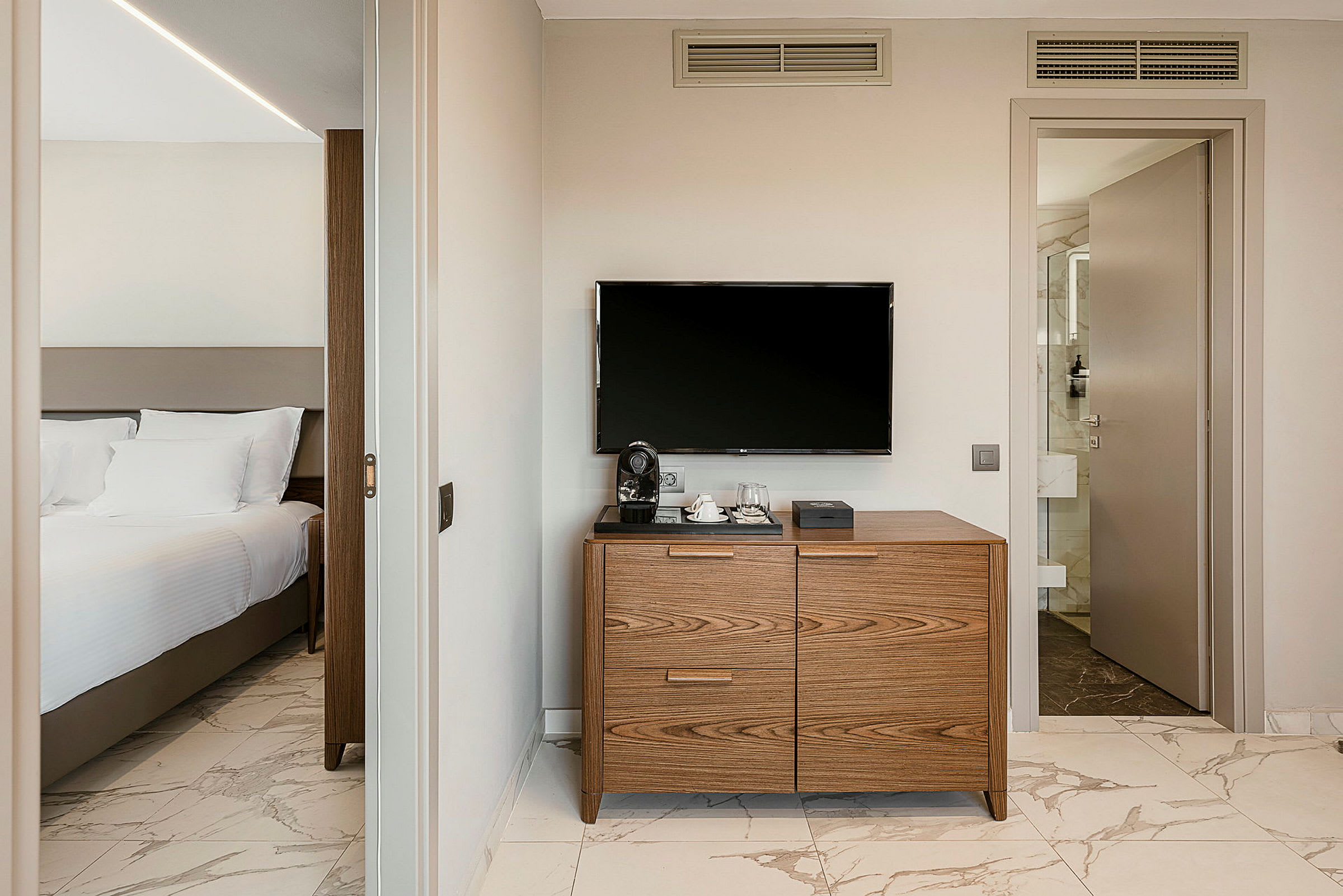 Et hotellrom med en seng med hvitt sengetøy til venstre. En kommode med en kaffemaskin står under en TV på veggen til høyre. (AI-generert tekst)