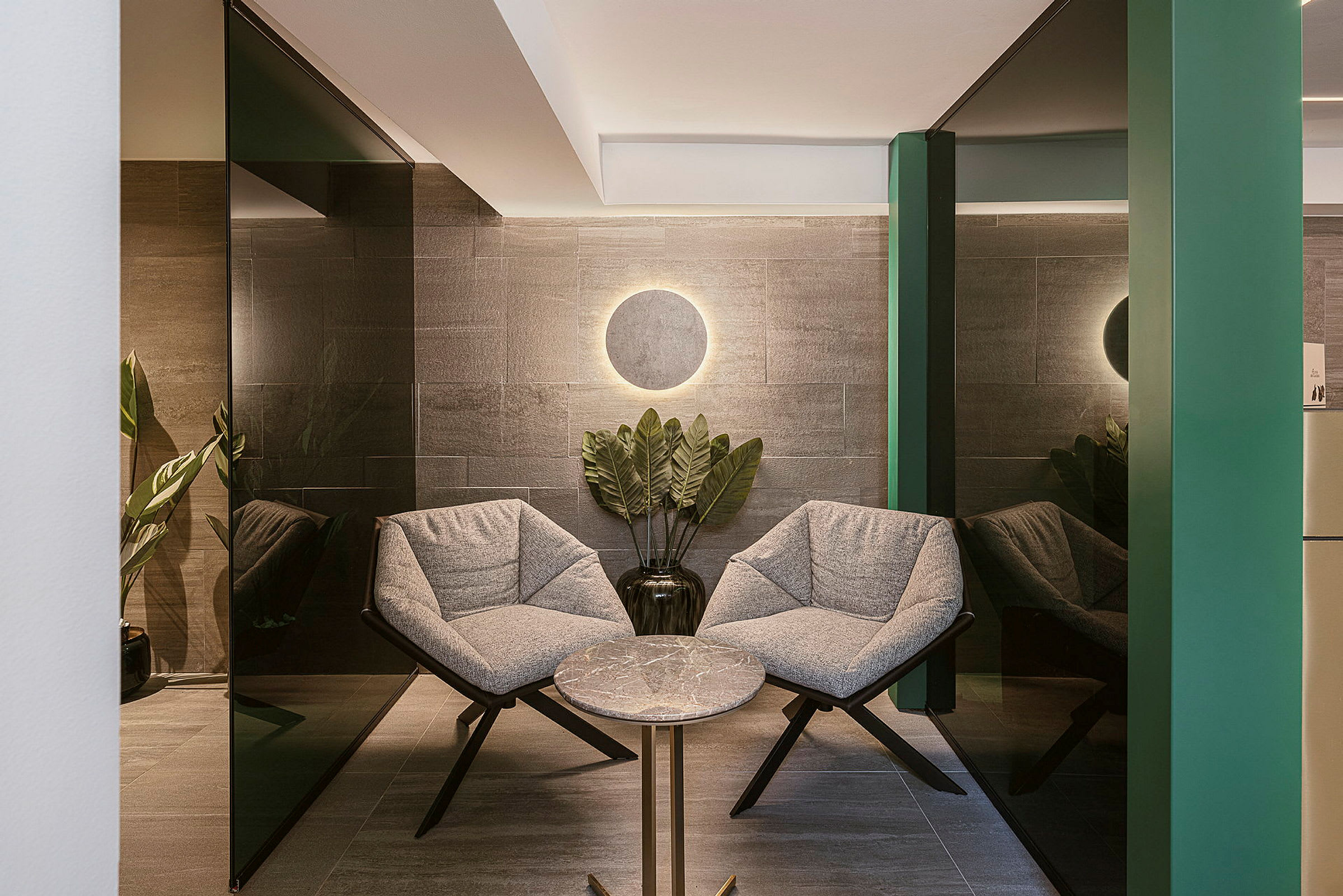 To grå lenestoler er plassert rundt et rundt bord i et moderne og elegant miljø. Bak møblene står en mørk vase med en grønn plante. (AI-generert tekst)