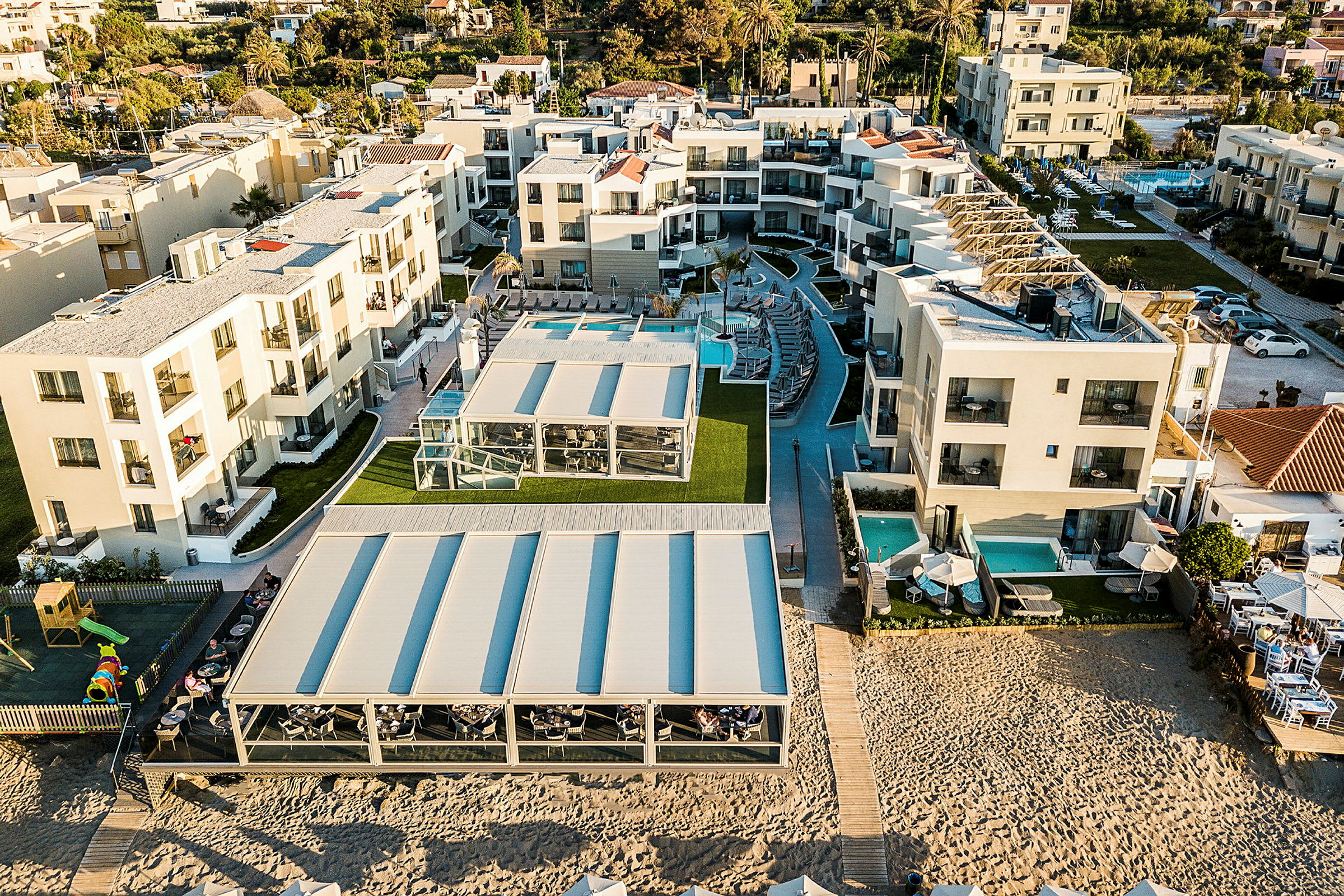 Luftbilde av et hotellområde med moderne bygninger, bassenger og uteområder nær sandstrand. (AI-generert tekst)