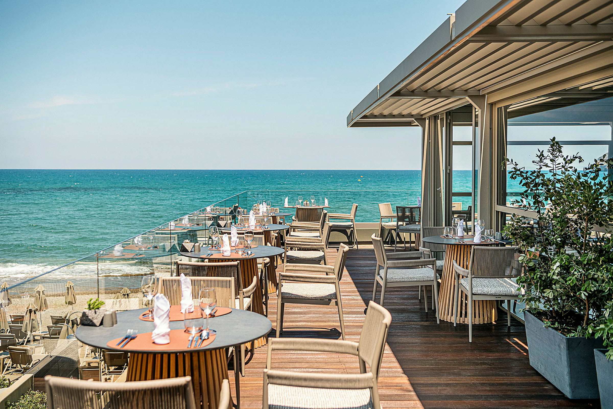 En terrasse med bord og stoler ved havet. Blått vann og klar himmel er synlig i bakgrunnen. (AI-generert tekst)