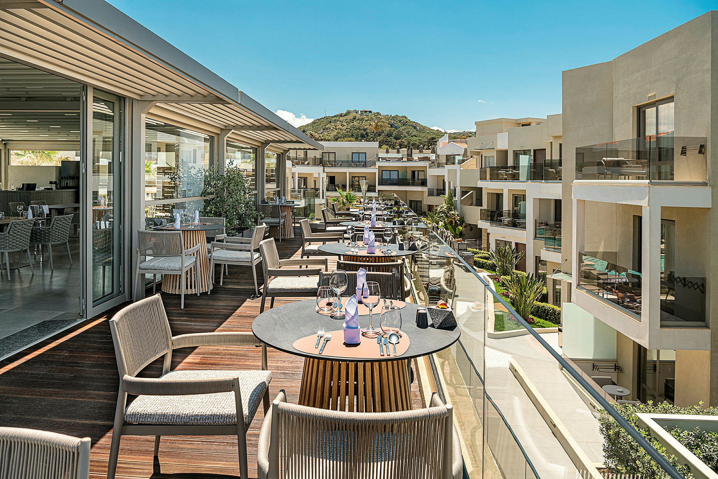 En terrasse med bord og stoler med utsikt over moderne bygninger og grønne omgivelser. (AI-generert tekst)