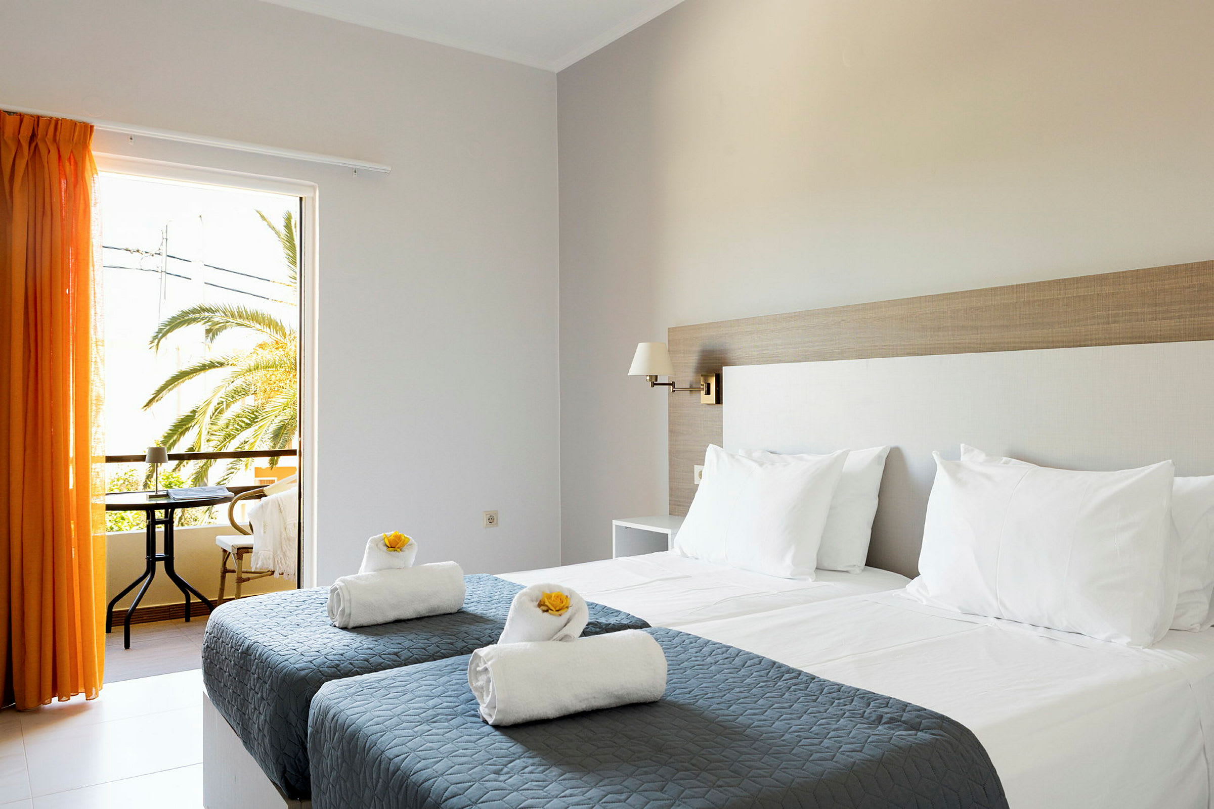 Et hotellrom med to enkeltsenger med grå sengetepper og hvite puter samt håndklær dekorert med gule blomster. Et vindu med oransje gardiner åpner ut til en balkong med utsikt over palmer. (AI-generert tekst)