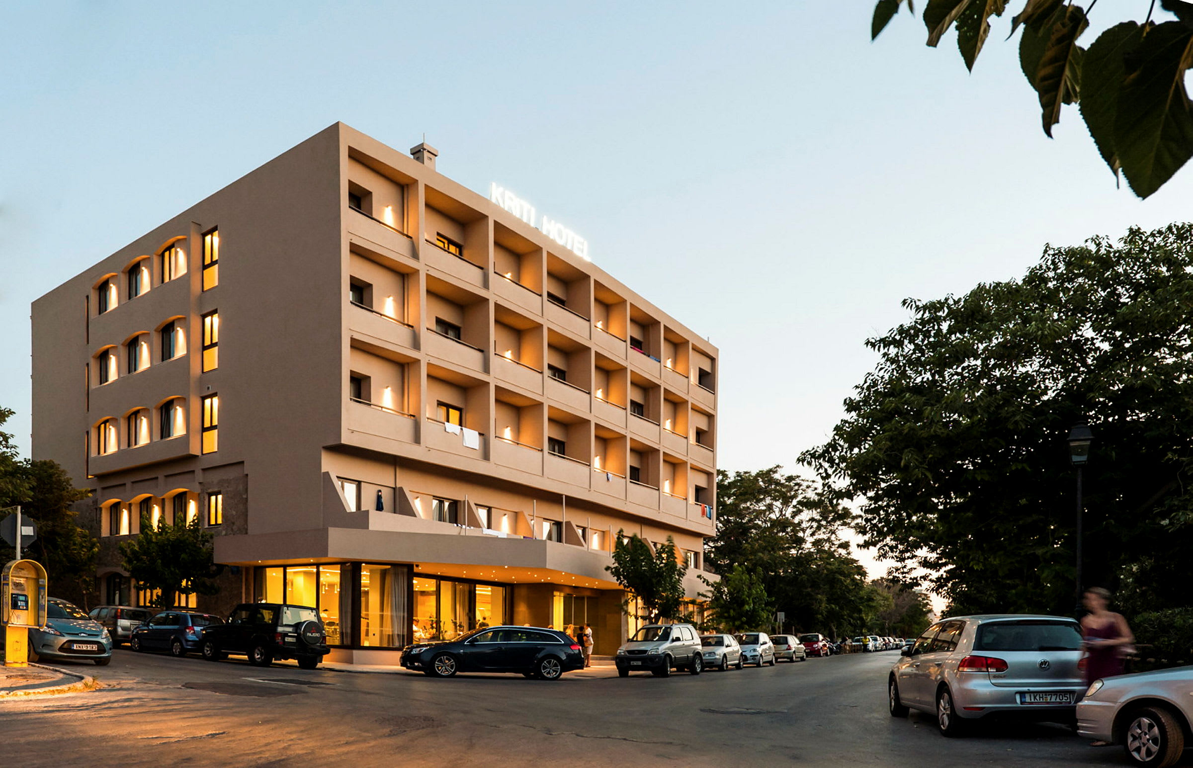 Fireetasjes hotellbygning med opplyst fasade, parkerte biler rundt og grønne trær i bakgrunnen. (AI-generert tekst)