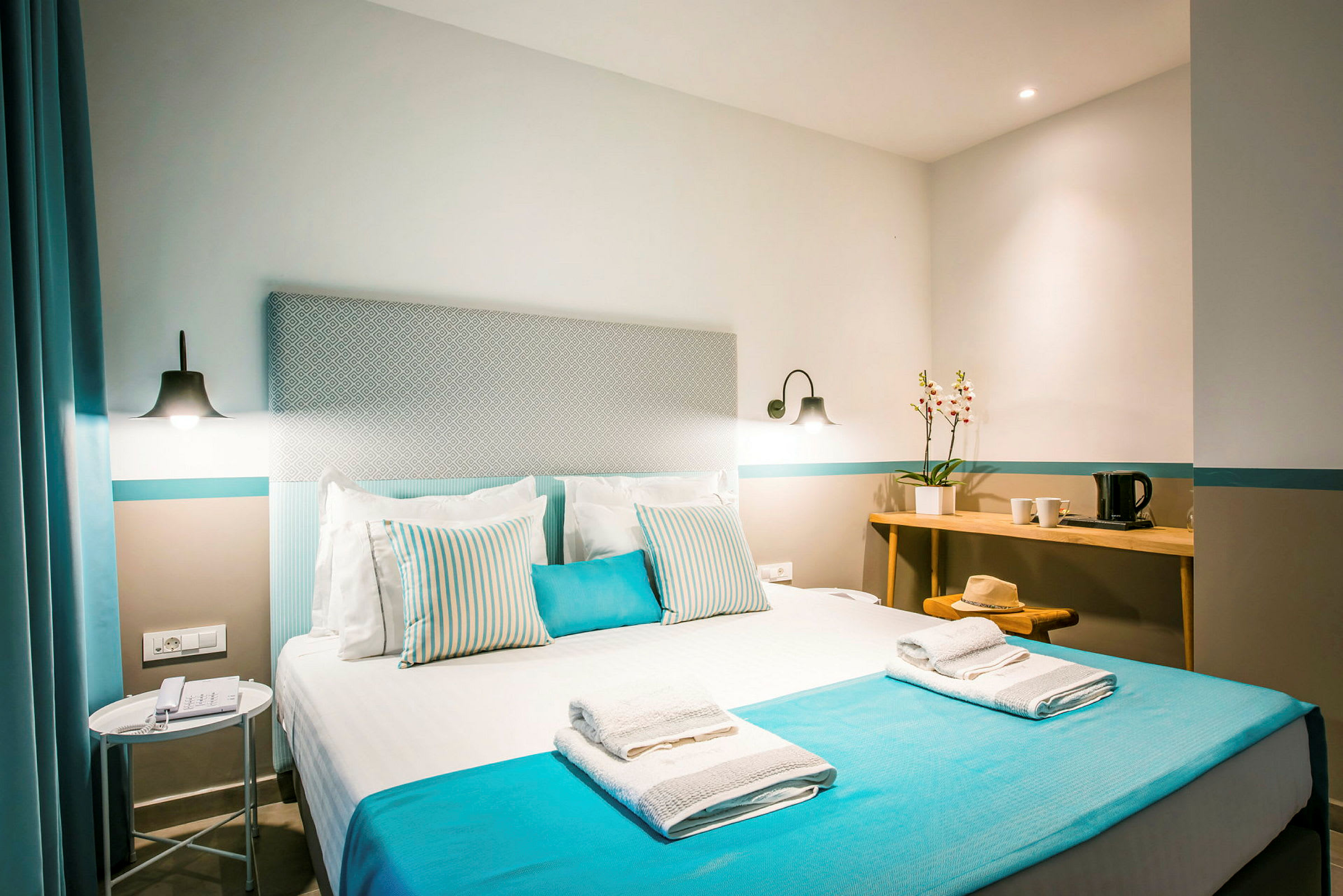 Lyst og moderne hotellrom med en dobbeltseng oppredd med blå og hvite detaljer. På sengen ligger håndklær, og det vises et sidebord med lampe samt et skrivebord med dekorativ blomst og vannkoker. (AI-generert tekst)