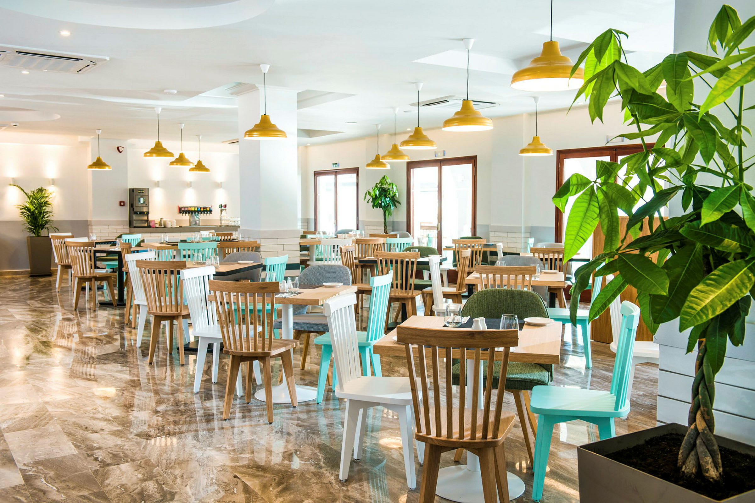 En restaurant med bord og stoler, samt store vinduer og grønne planter. (AI-generert tekst)