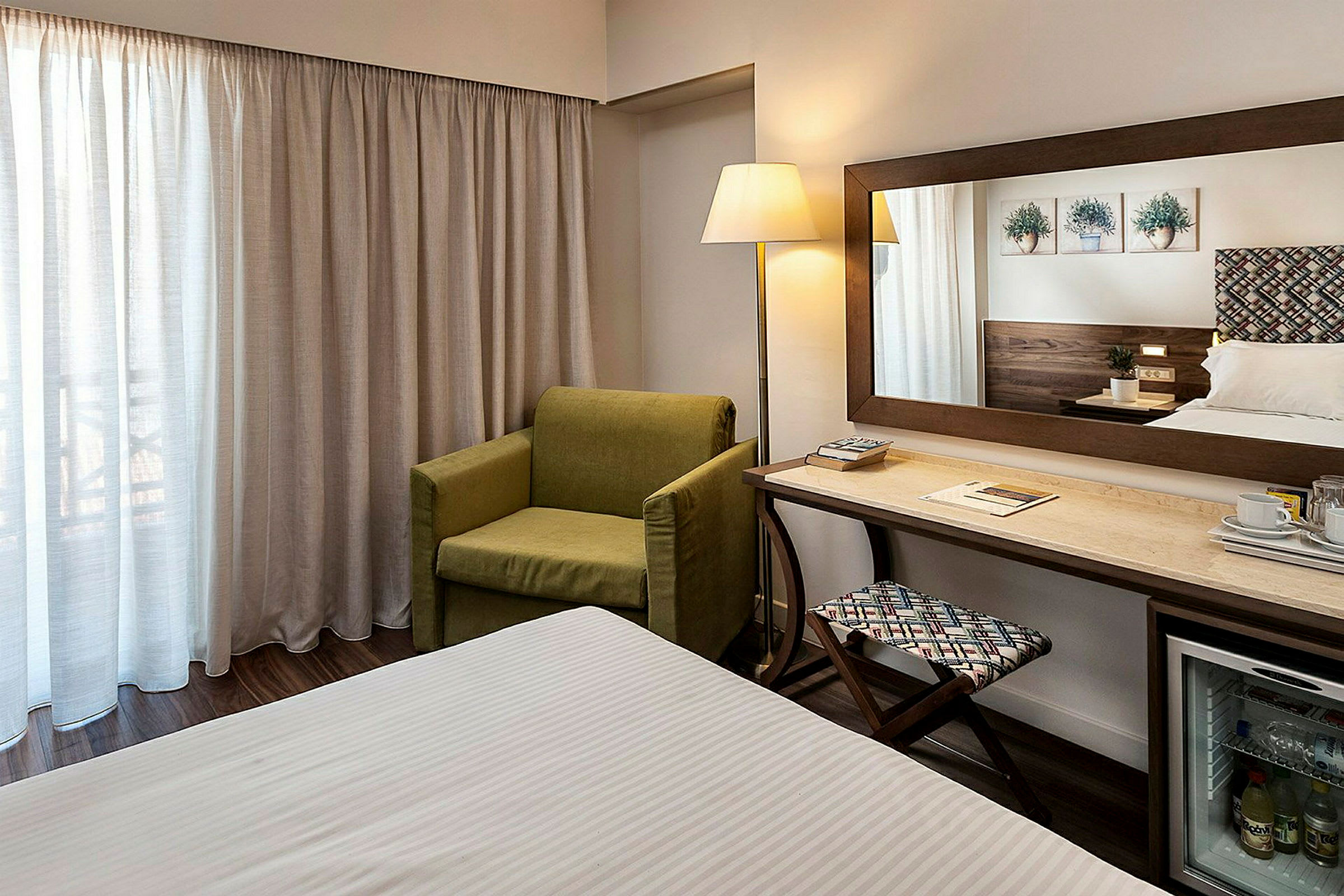 Hotellrom med grønn lenestol, skrivebord, speil og seng. (AI-generert tekst)