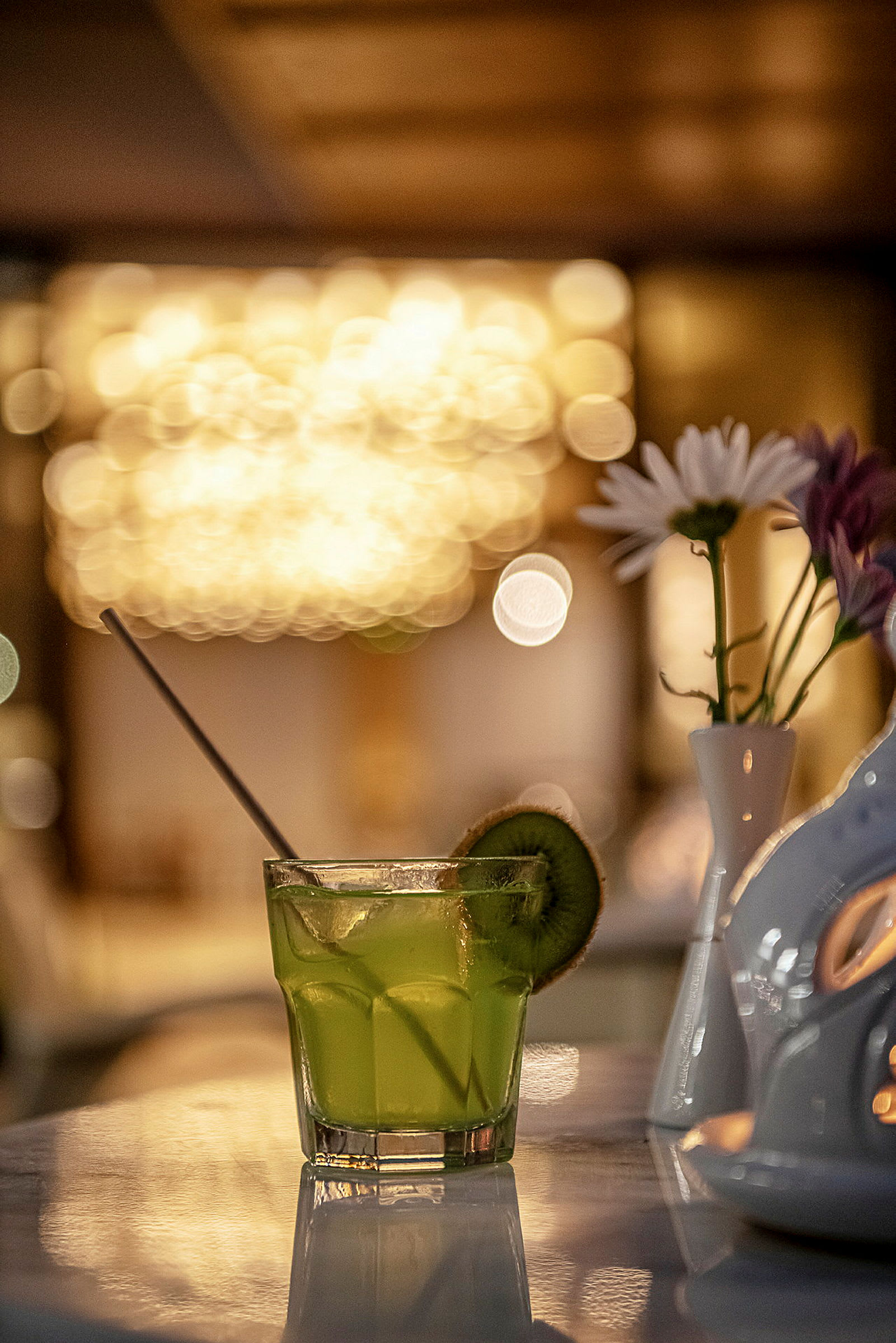 Et glass med en grønn drink dekorert med en kiwiskive på en marmorbord med en vase som inneholder blomster i bakgrunnen. (AI-generert tekst)