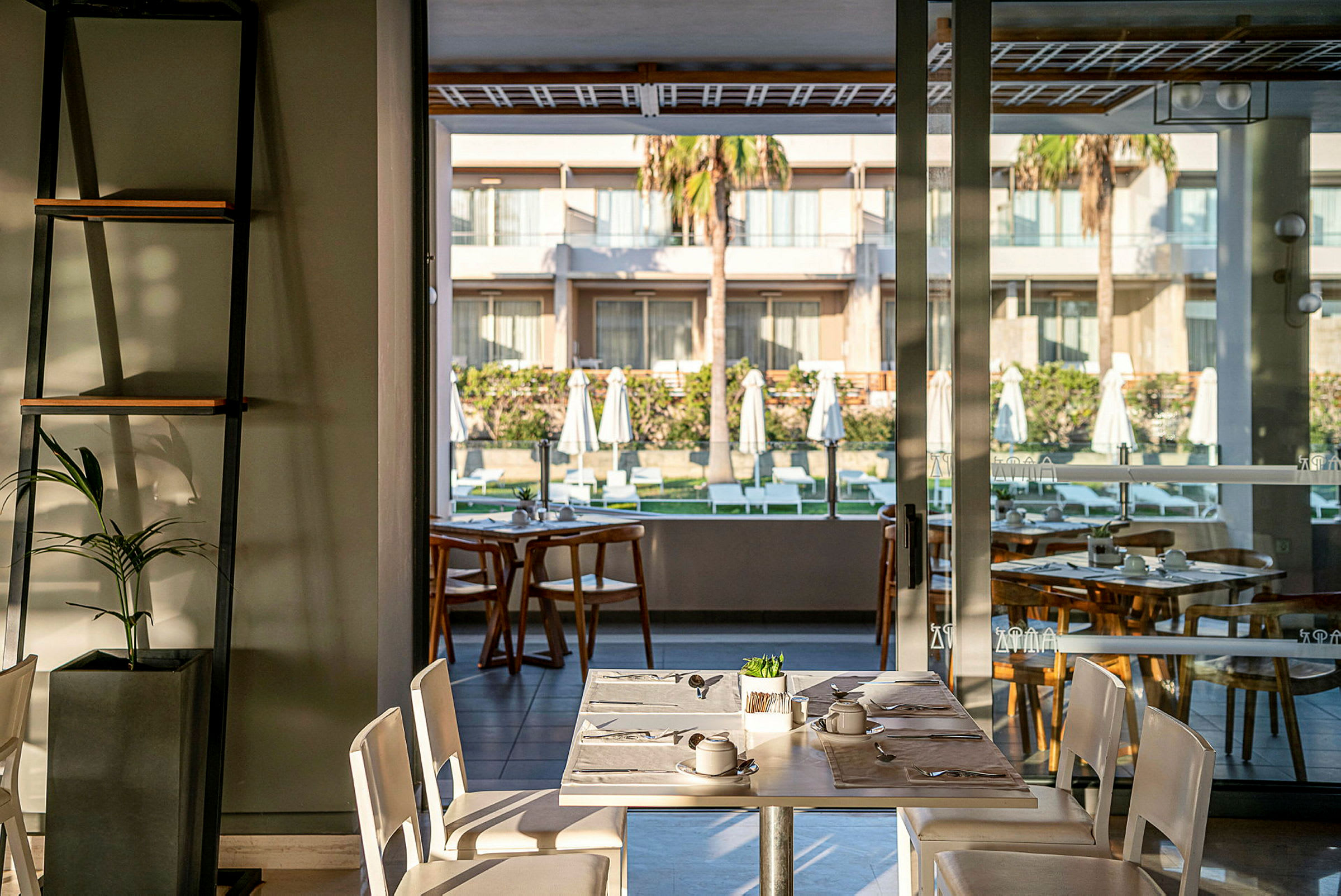 En restaurant med bord i forgrunnen og utsikt over en indre gårdsplass med solsenger og parasoller. (AI-generert tekst)