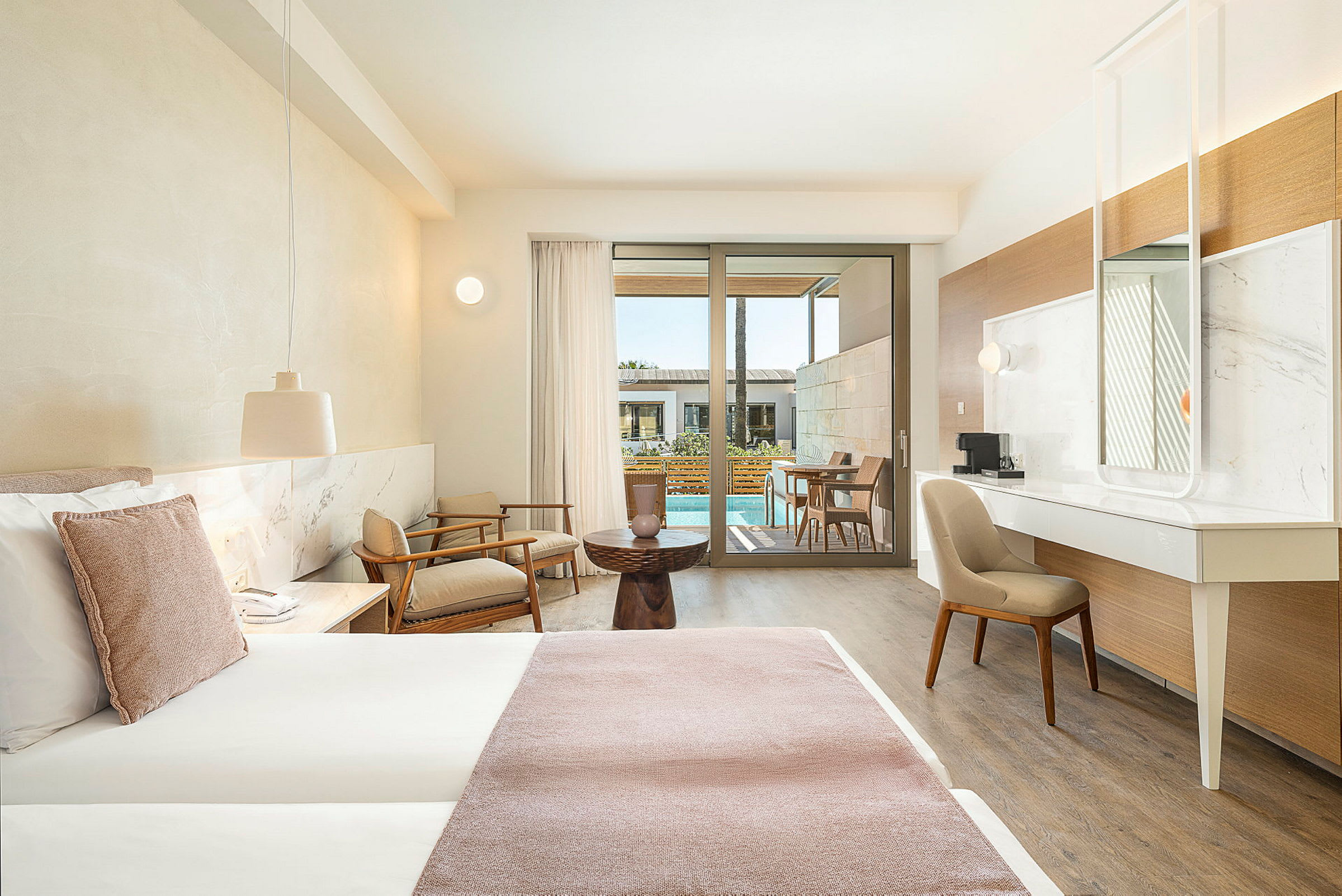 Et hotellrom med moderne innredning, en stor seng, sittegruppe og utsikt mot en terrasse med basseng. (AI-generert tekst)