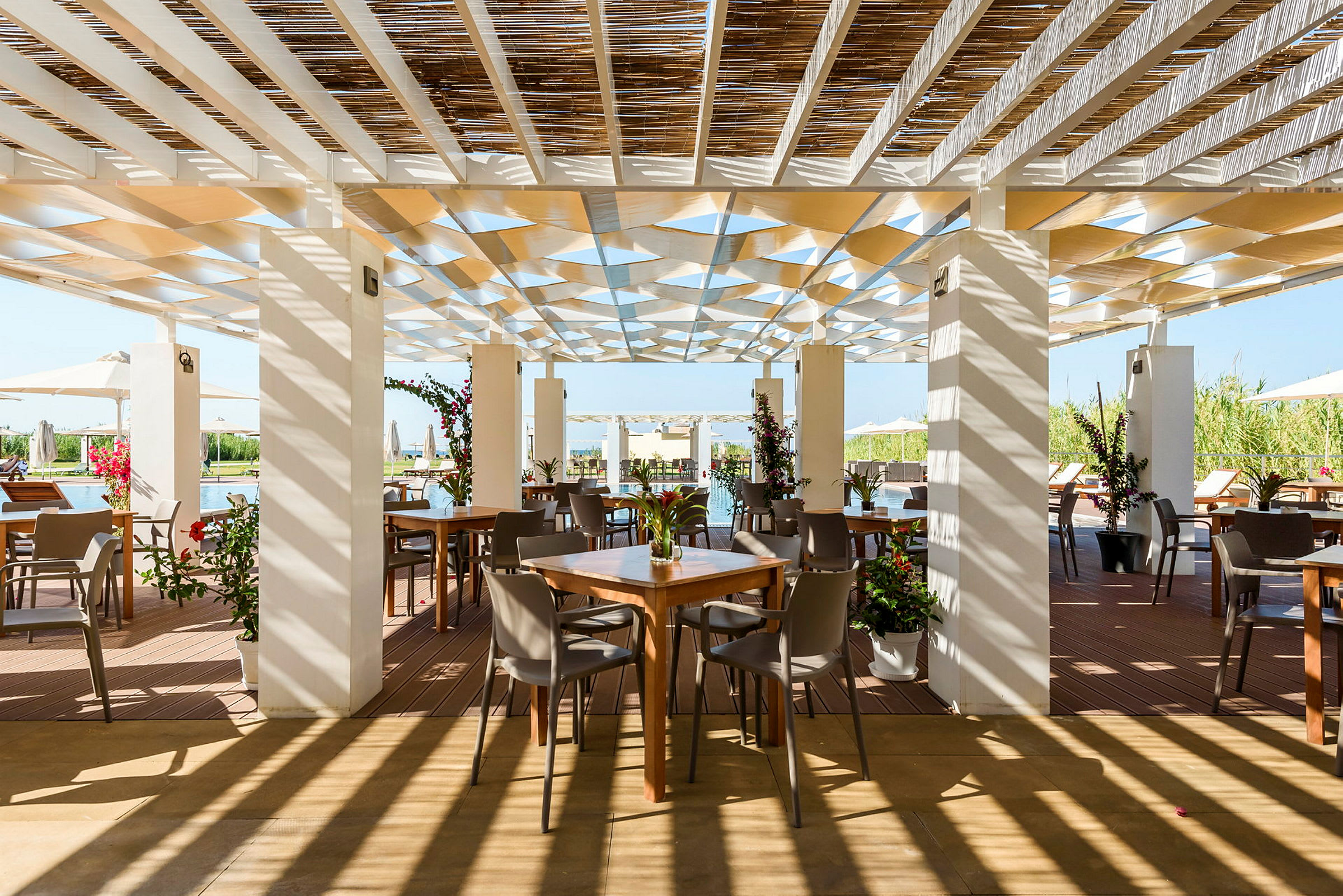 En utendørs restaurant med bord og stoler under en skyggefull pergola, omgitt av grøntområder. (AI-generert tekst)