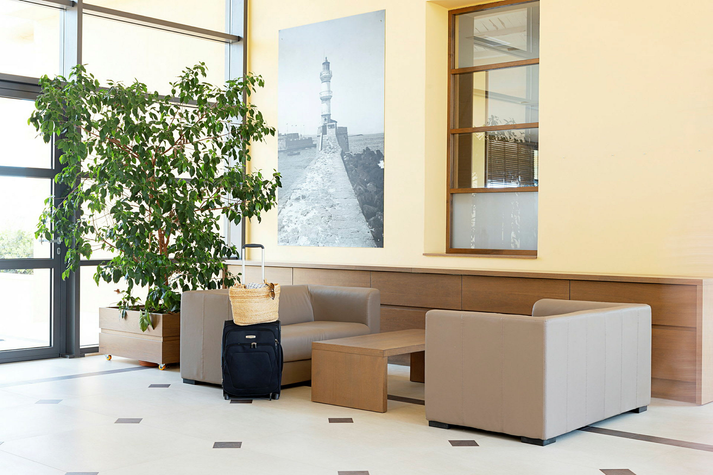 En resepsjonslounge med beige lenestoler, et bord, en stor grønn plante og en svart koffert med en kurv oppå. (AI-generert tekst)