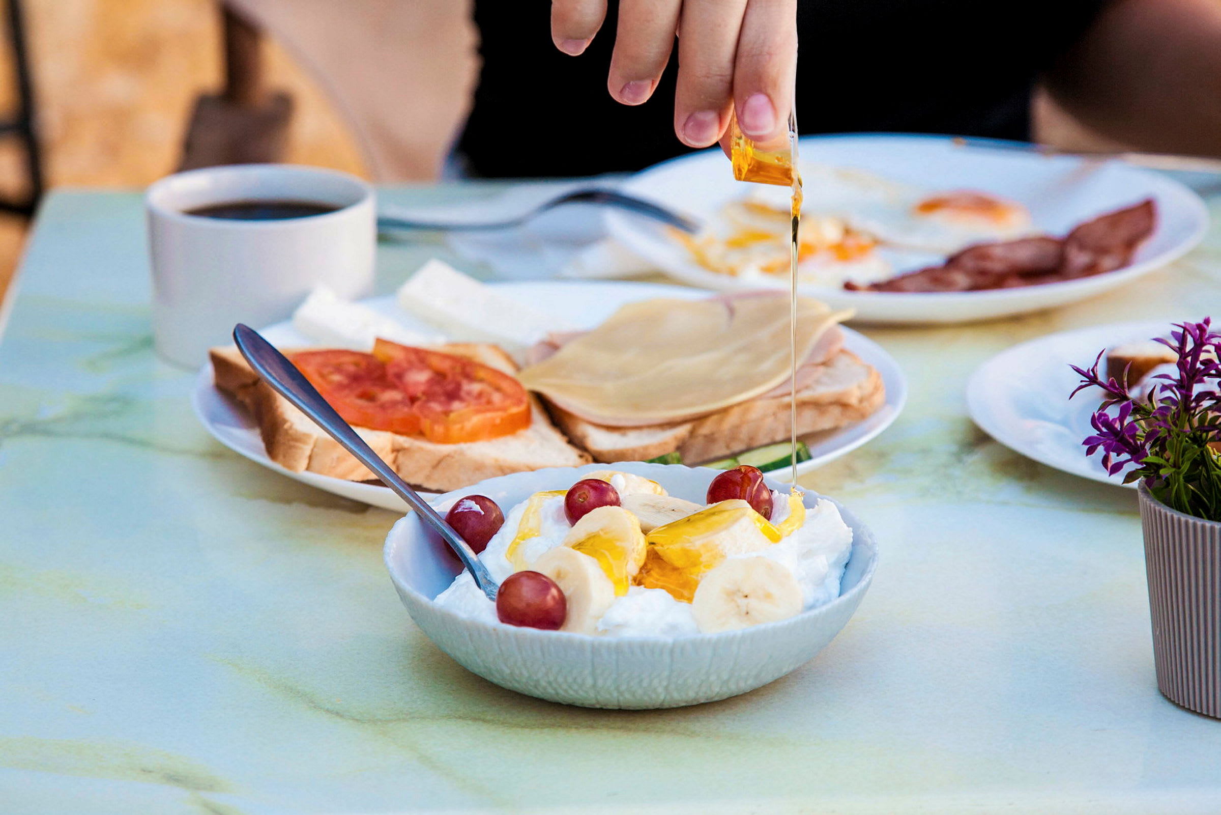 En hånd heller honning over en skål med yoghurt, banan og druer. I bakgrunnen er det en kaffekopp og tallerkener med brød og pålegg. (AI-generert tekst)